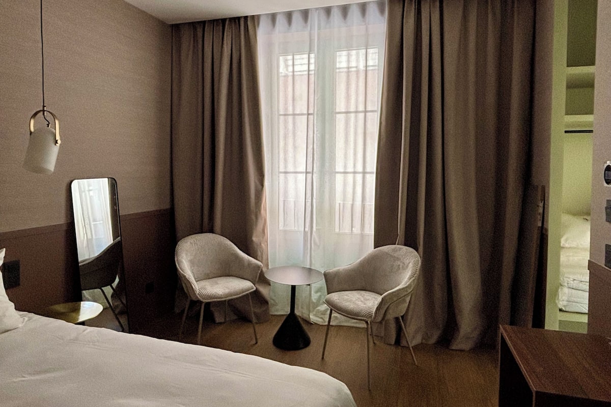 A hotel room with a bed, two chairs, a small table, a mirror, and a window with curtains.