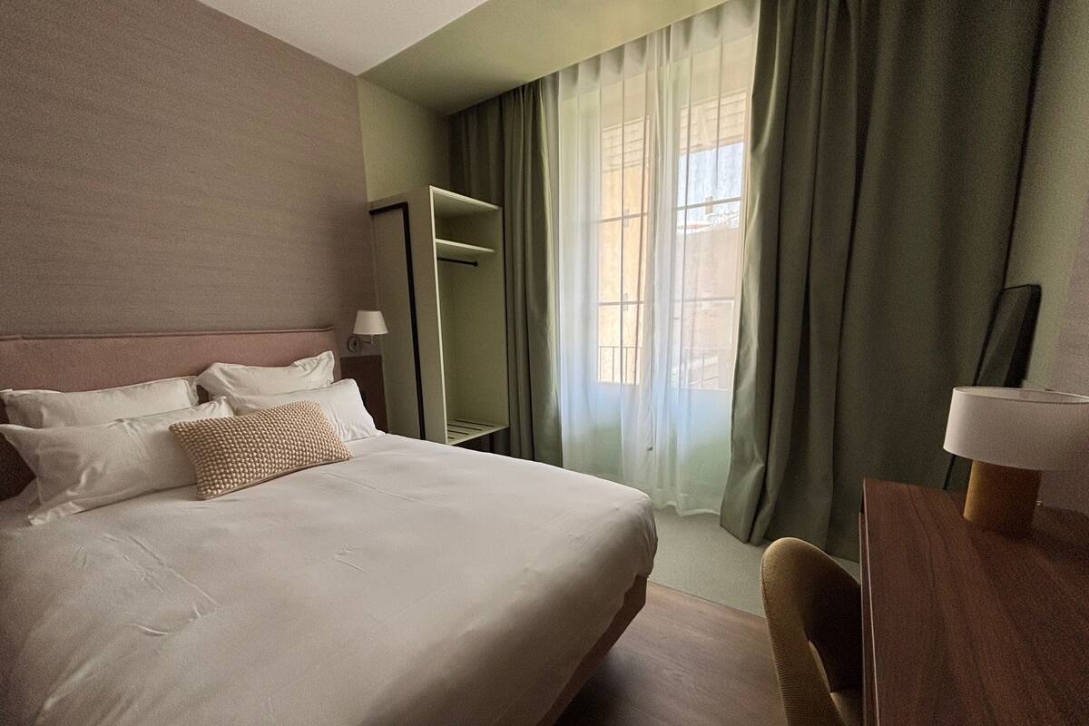 A hotel room with a bed, a desk, a chair, a wardrobe, and a window with curtains.