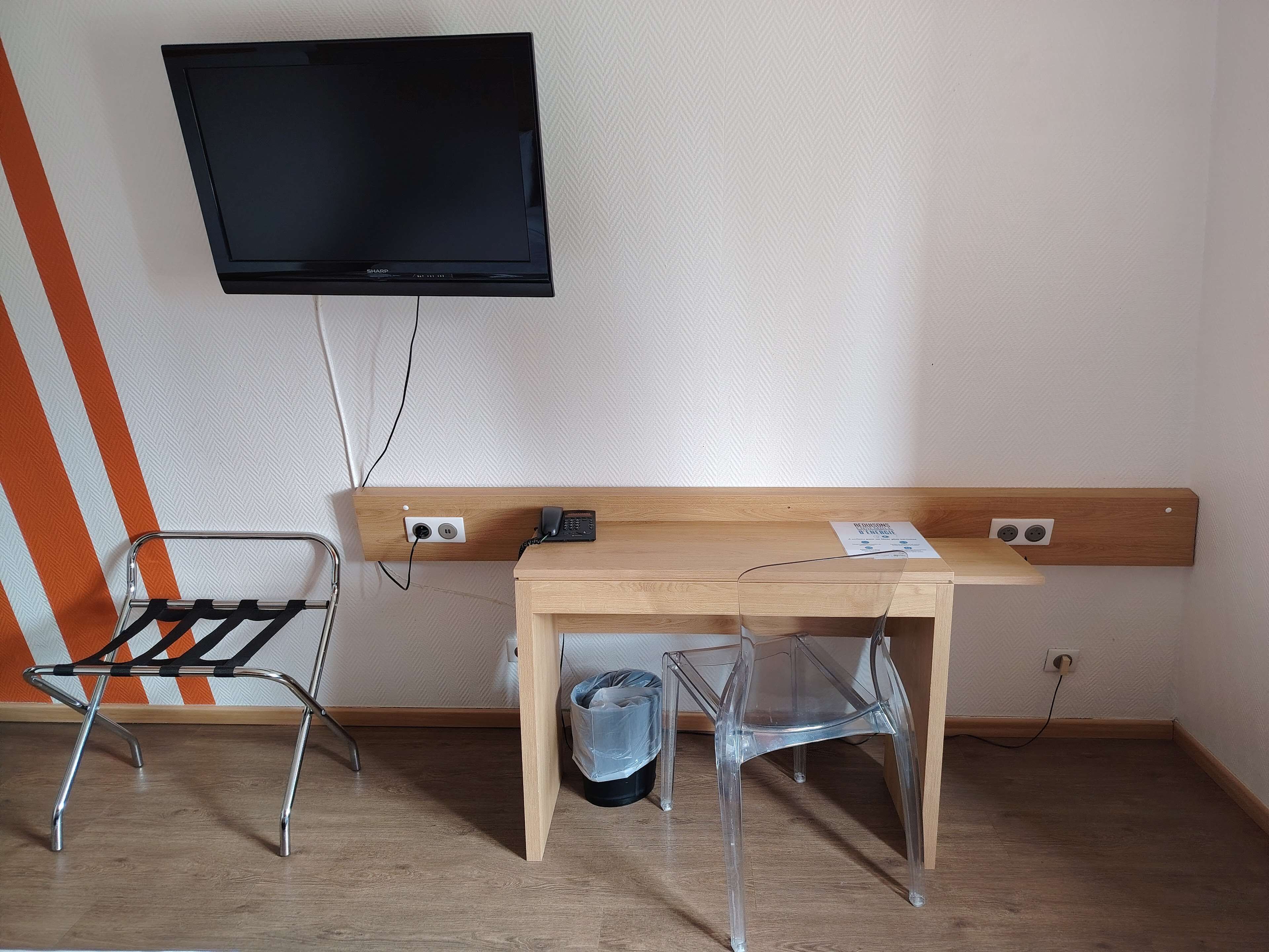 A room with a wooden desk, a chair, a television mounted on the wall, and a trash bin.