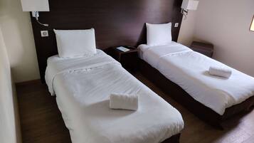 A hotel room with two single beds, each with white linens and a rolled towel. There are bedside tables with lamps and a nightstand.