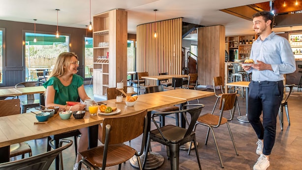 A modern restaurant with wooden tables and chairs, a bar area, and a visible outdoor seating area. - Kyriad Laon (Laon)