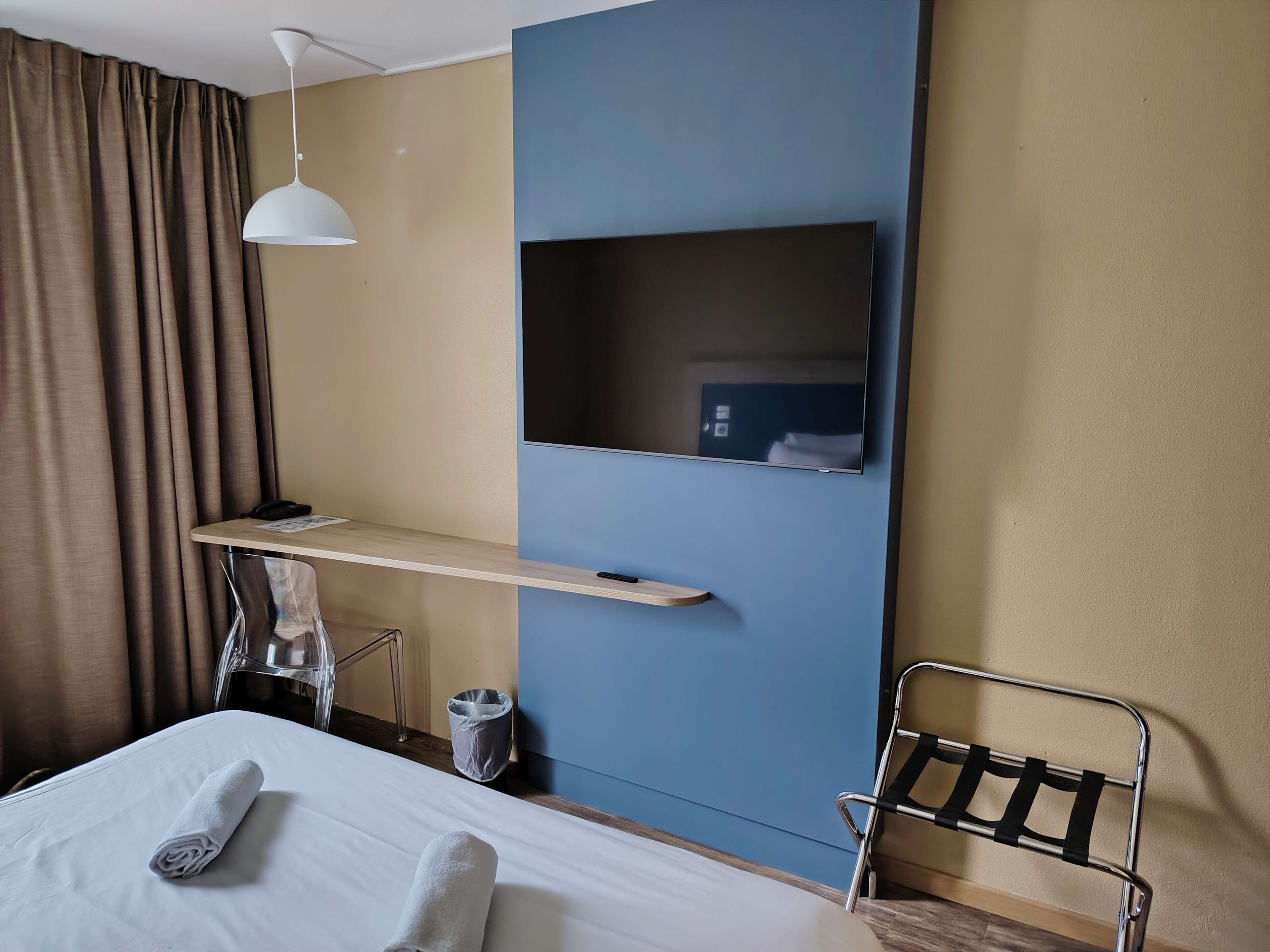 Habitación de hotel con cama, escritorio, silla, televisor y lámpara colgante.