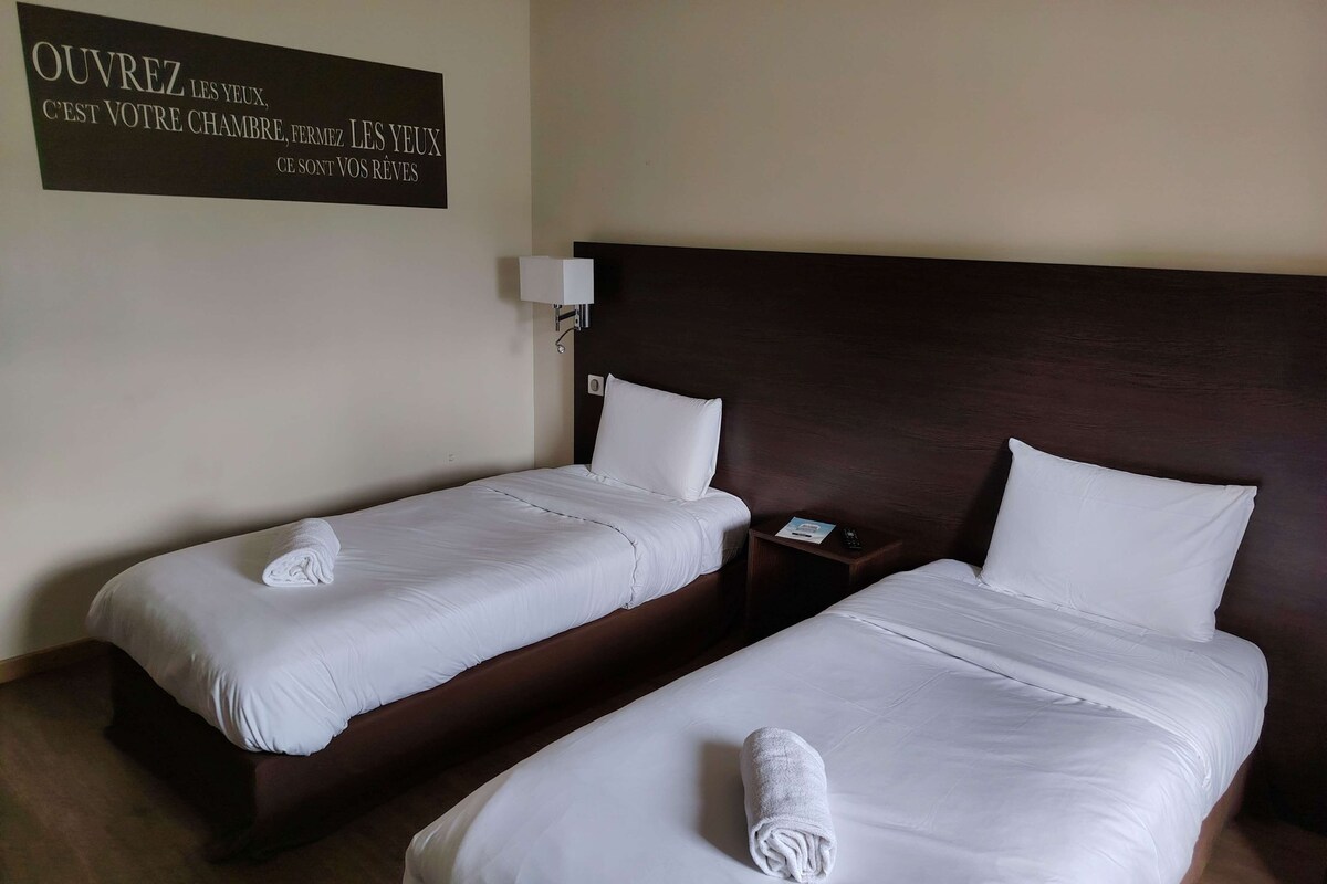 A hotel room with two single beds, each with a white duvet and a rolled towel. There is a dark wooden headboard, a bedside table with a lamp, and a wall sign with text.
