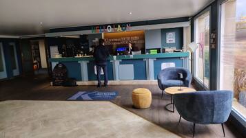 Een hotel receptie met een balie, twee stoelen, een klein rond tafeltje en een groot raam.