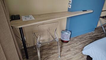 A small room with a wooden desk, a clear plastic chair, a trash bin, and a bed with a white cover.