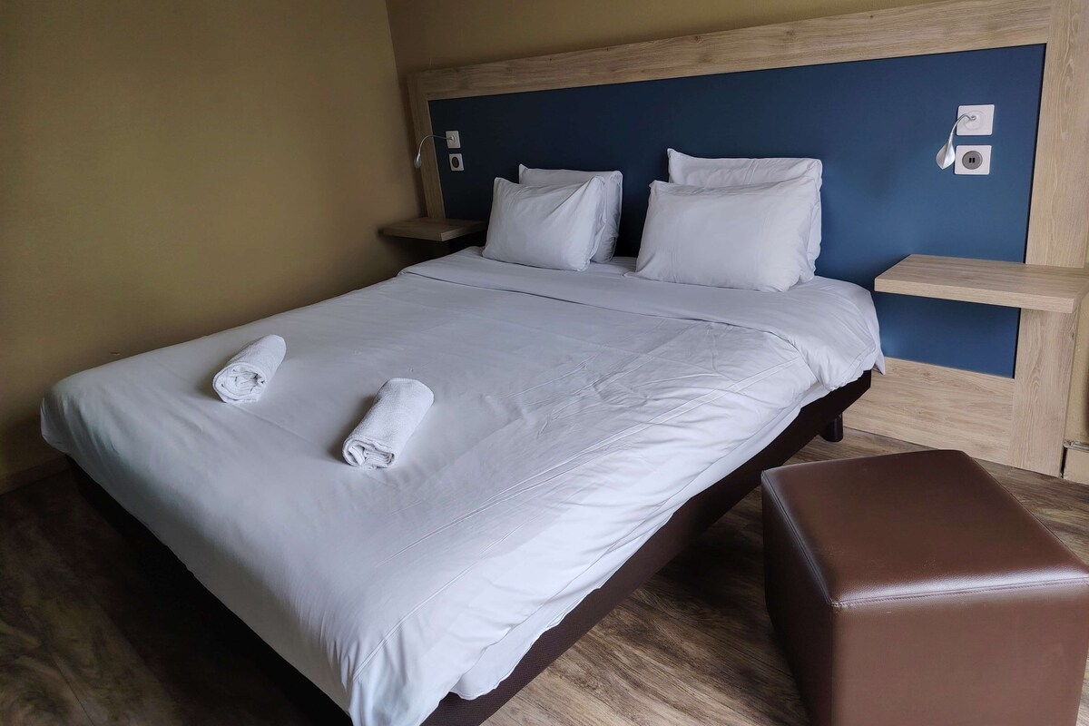 A neatly made bed with white linens and two rolled towels, a wooden headboard, and a brown bench.