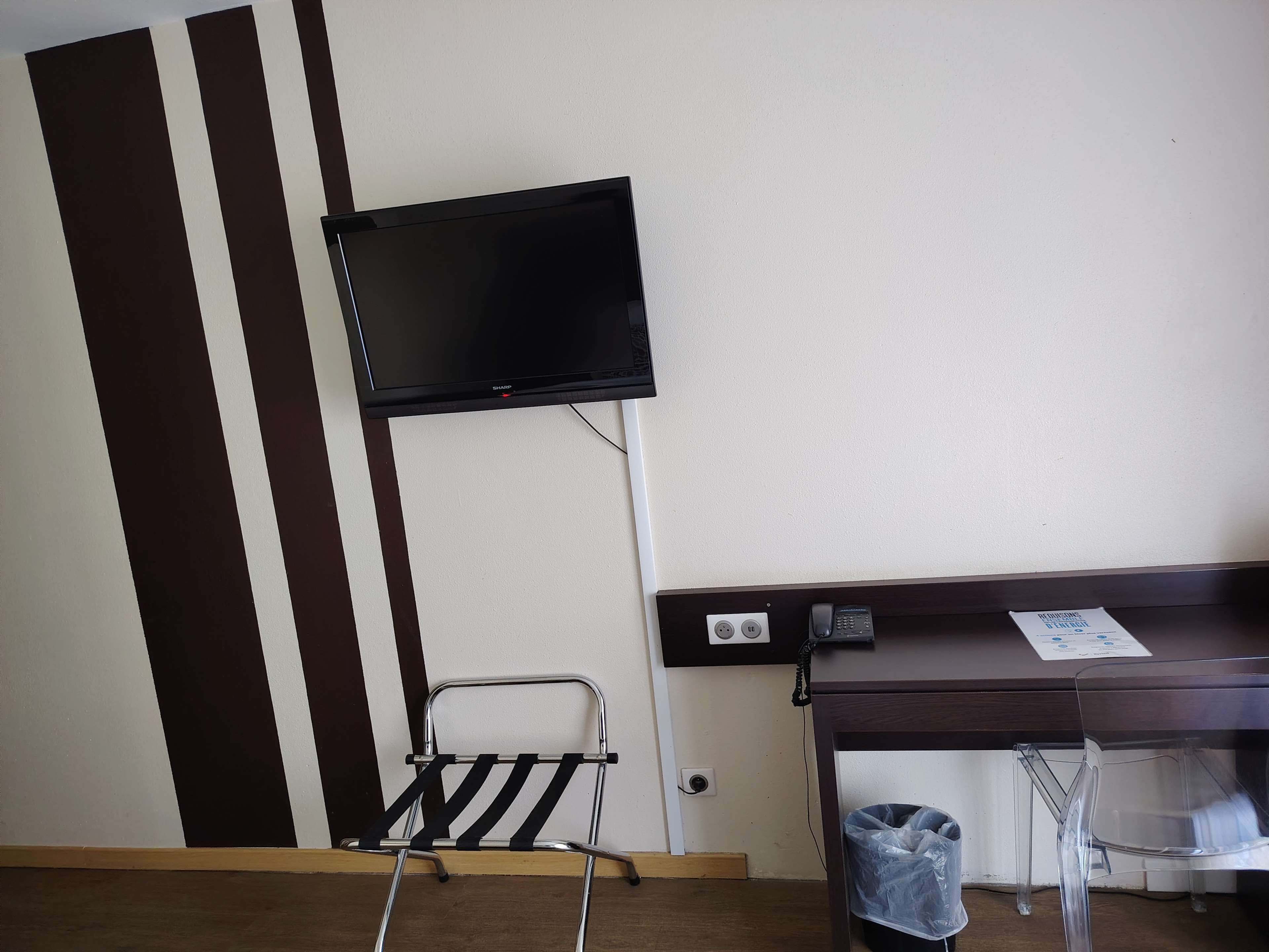 A hotel room with a flat-screen TV mounted on the wall, a desk with a telephone and a paper, and a chair with a black seat.