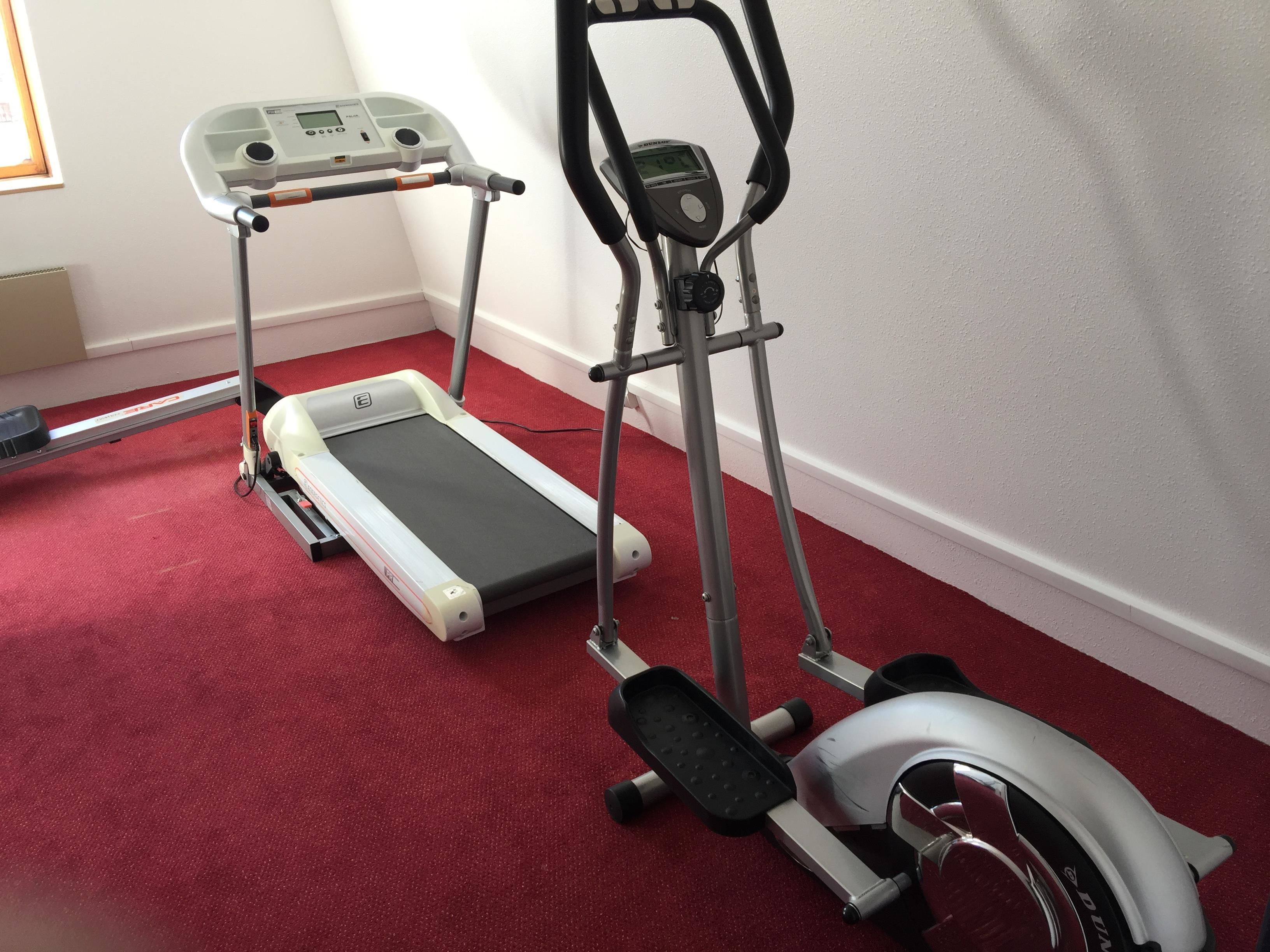 Twee stilstaande fitnessapparaten op een rood tapijt in een kamer met witte muren.
