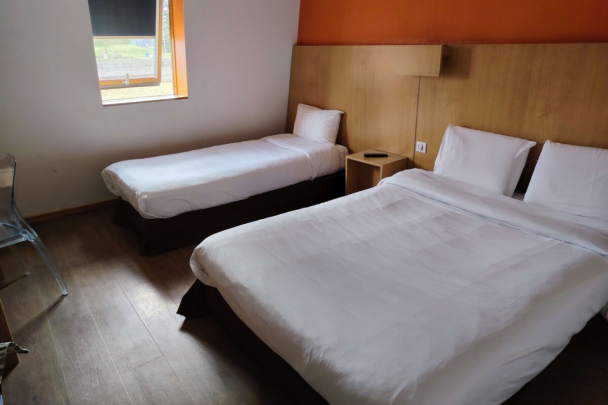 A hotel room with two single beds and one double bed, wooden headboard, a small bedside table with a remote control, and a window with a view of the outside.