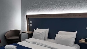 A hotel room with a large bed, two pillows, a blue blanket, a chair, a nightstand, and a wall-mounted light. - Kyriad Laon (Laon)