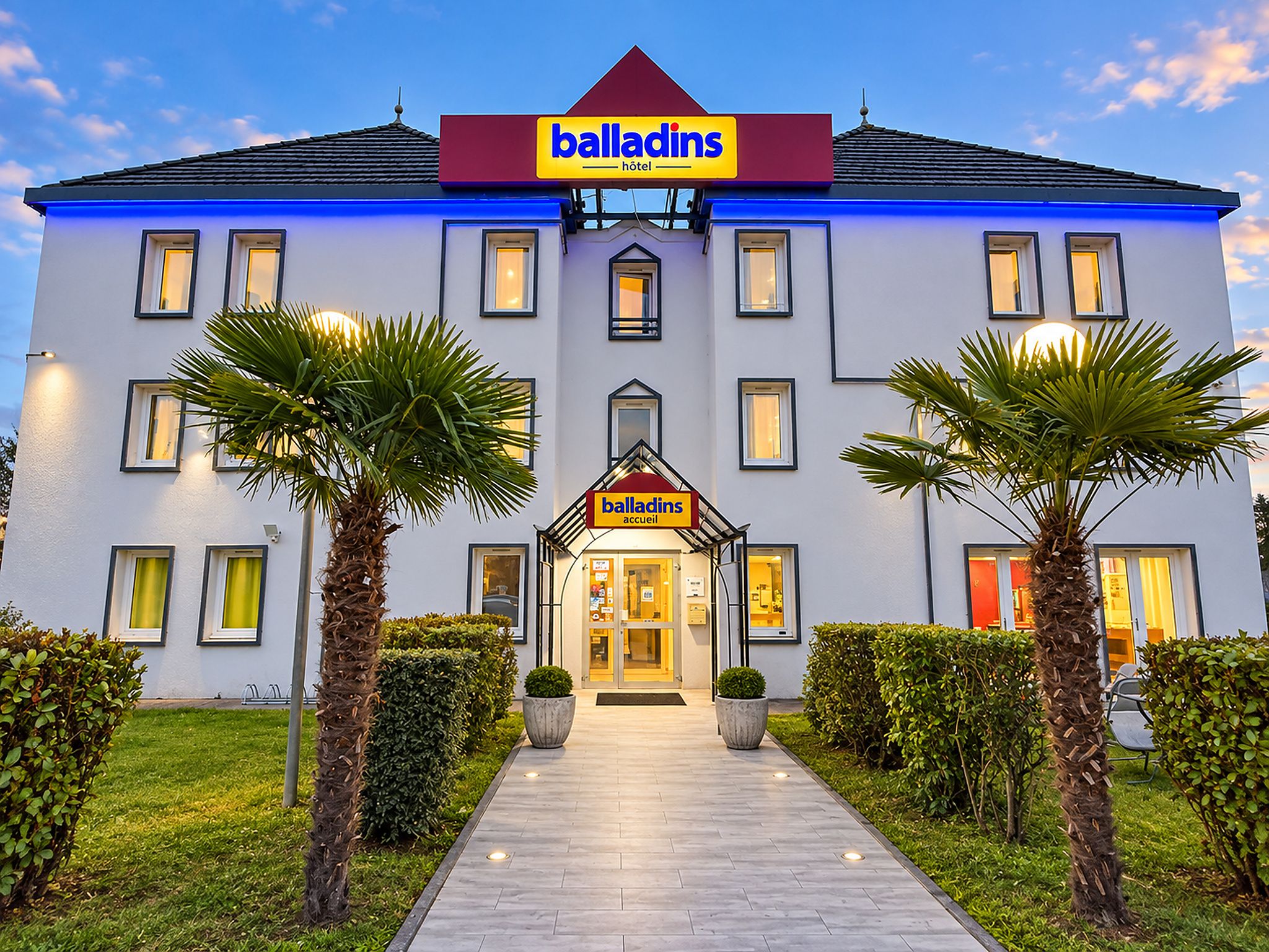 A hotel named Balladins with a modern facade, a central entrance, and symmetrical landscaping with palm trees.