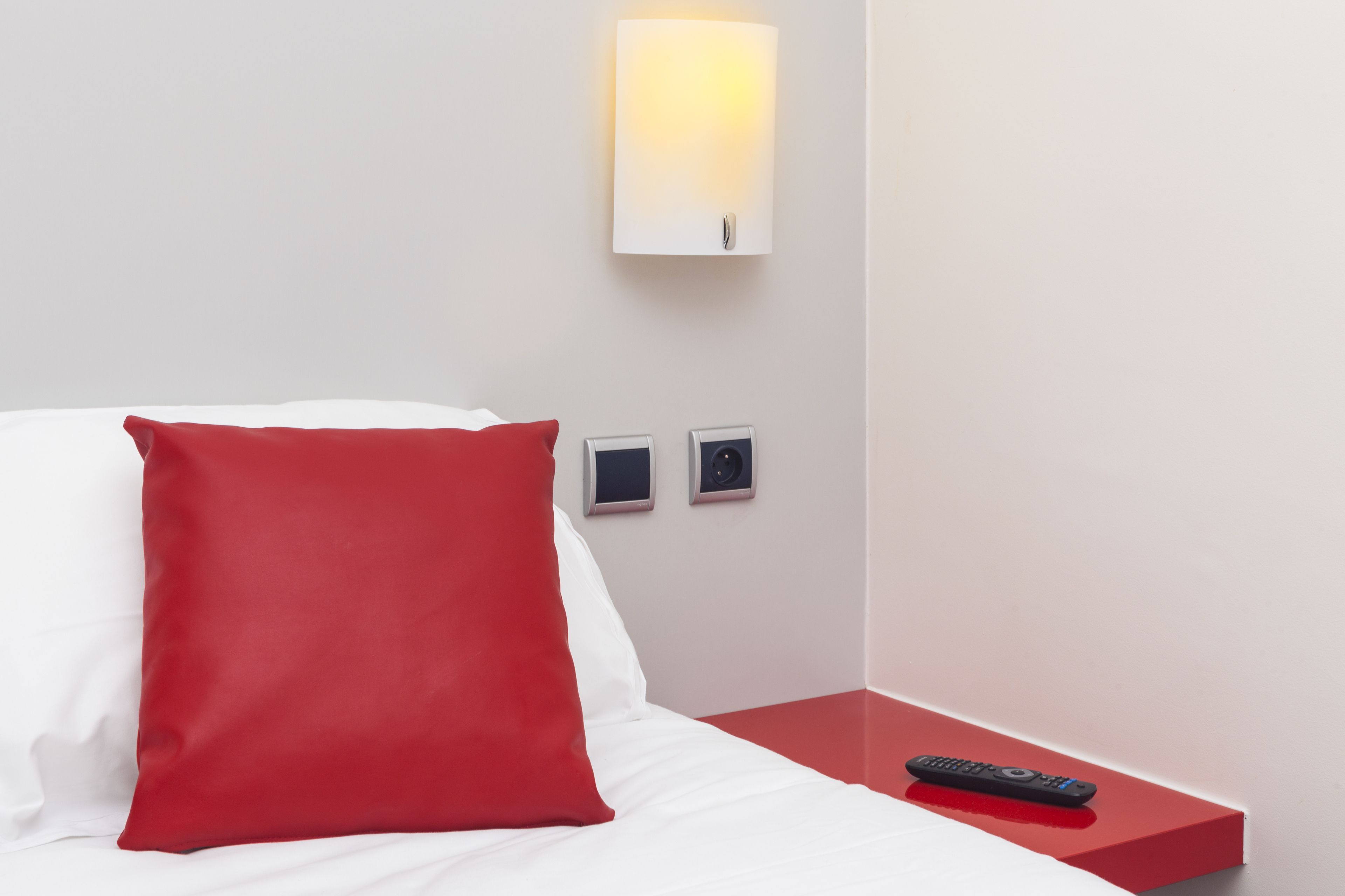 Hotellihuone, jossa on sänky, punainen tyyny, yöpöytä, jossa on kaukosäädin, ja seinälle asennettu valaisin.