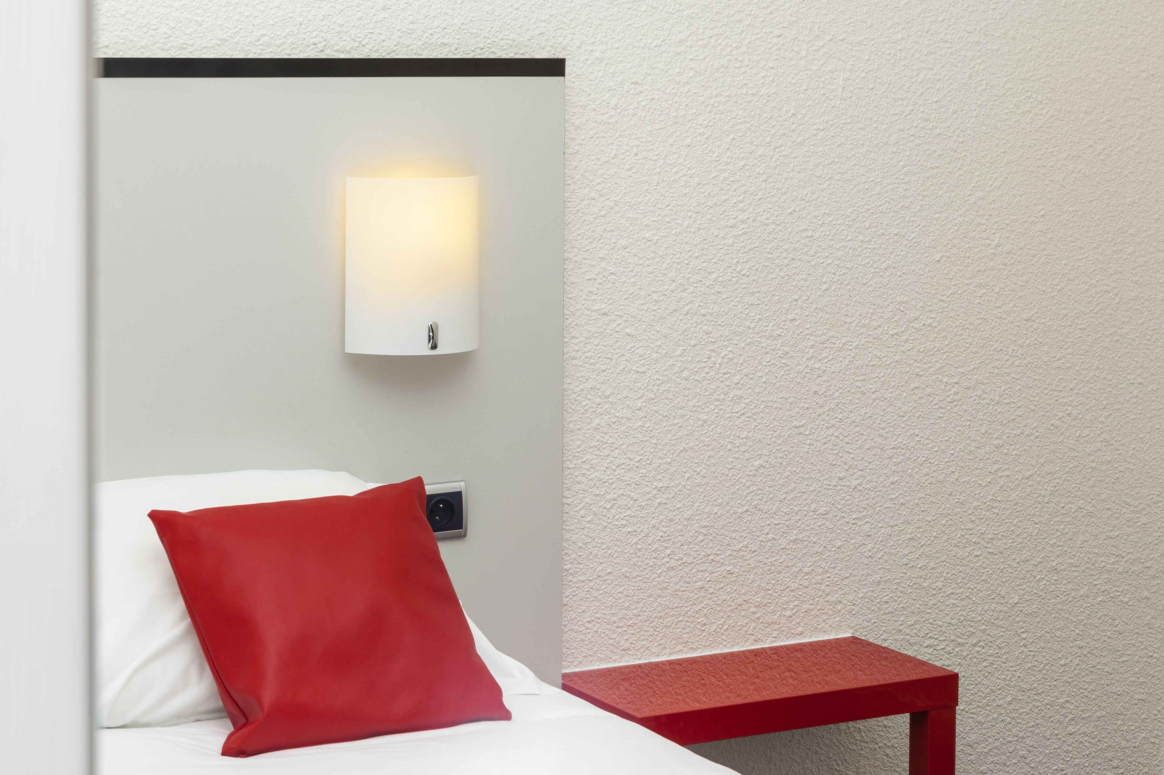 Et hotelværelse med en seng, en rød pude, et natbord, en vægmonteret lampe og et overvågningskamera.