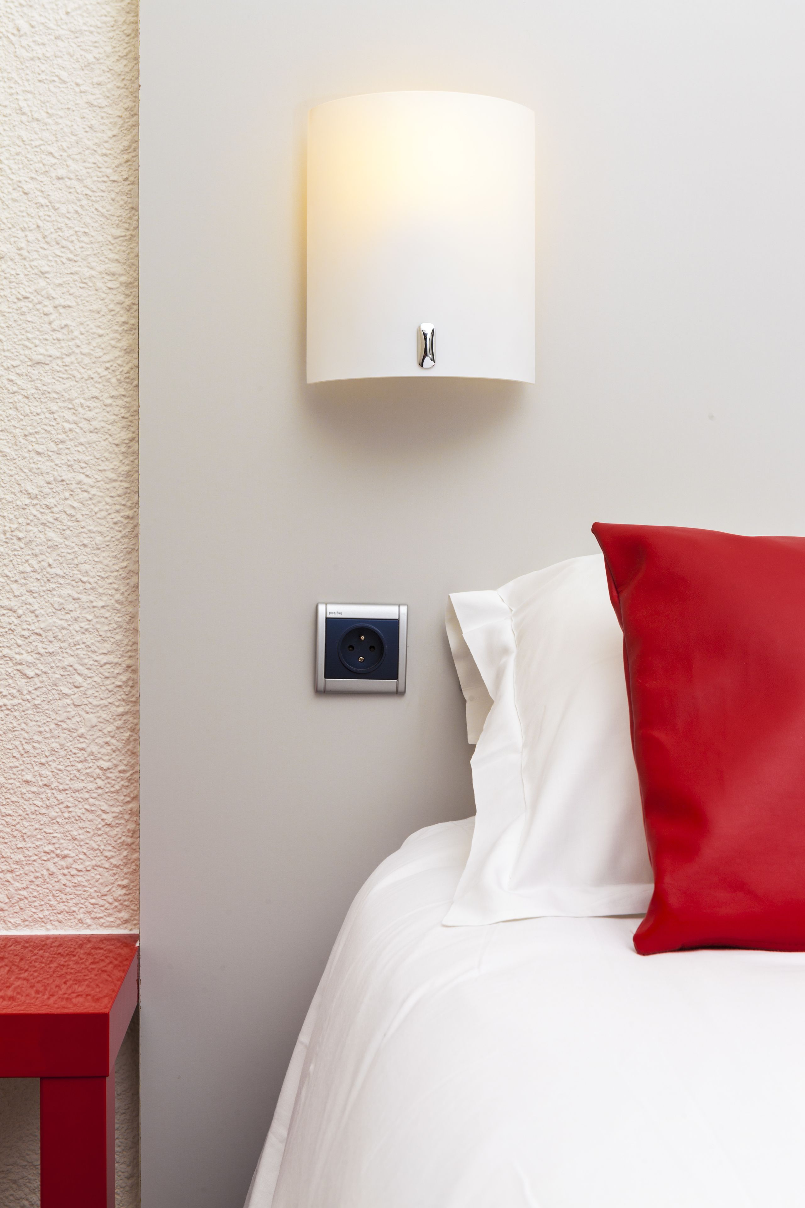 A hotel room with a bed, a red pillow, a wall-mounted light fixture, and a power outlet.