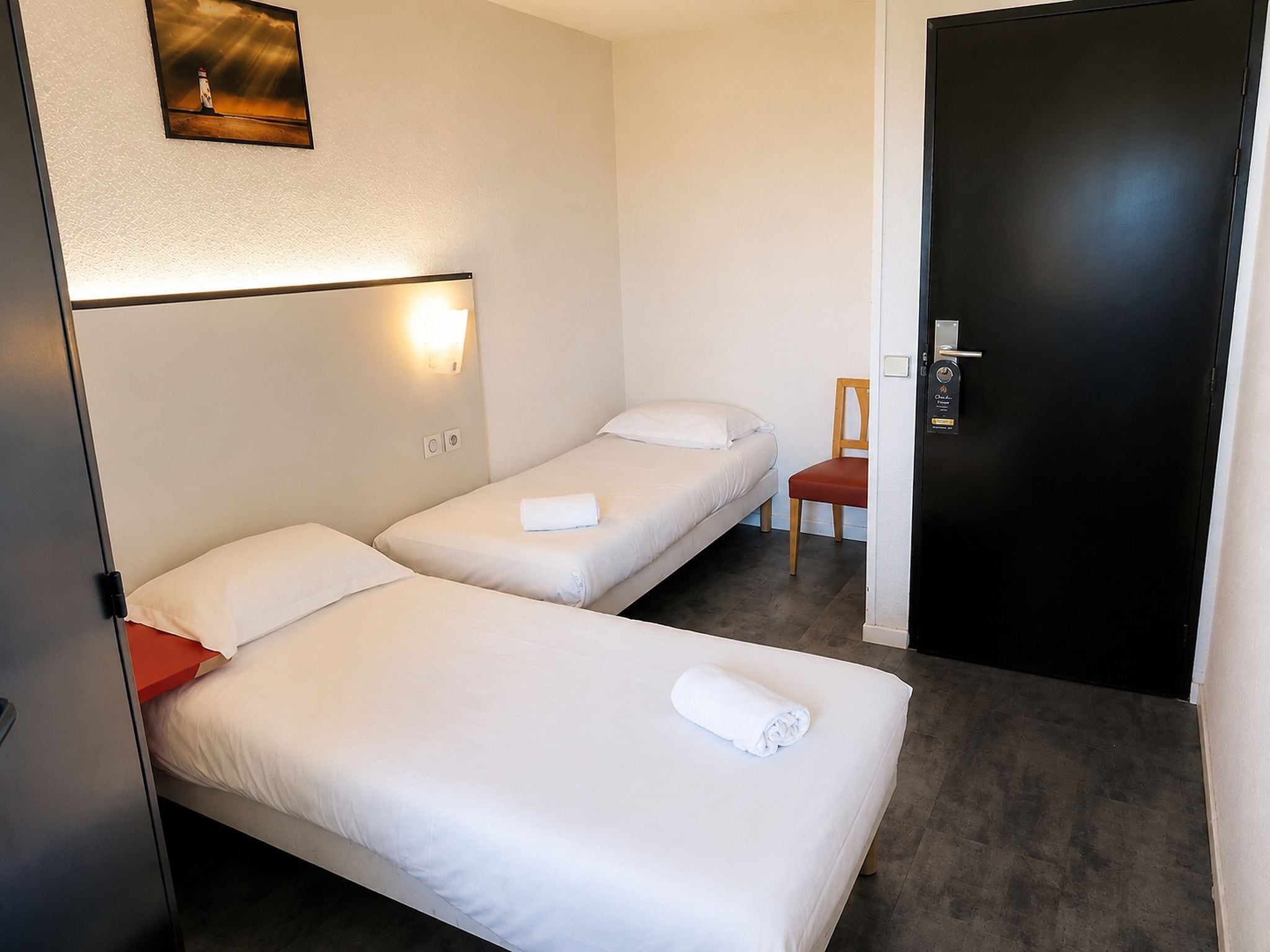 A hotel room with two single beds, a nightstand, a chair, and a door with a keycard lock.