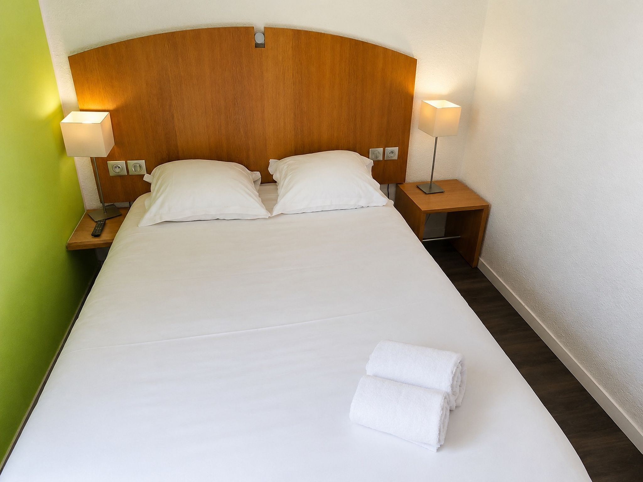 Hotellihuone, jossa on suuri sänky, kaksi yöpöytälamppua, puinen sängynpääty ja siististi pedattu sänky, jonka päällä on taiteltu pyyhe.