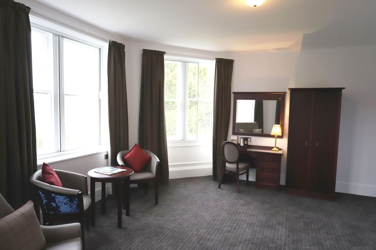 A hotel room with a large window, a desk with a lamp, a chair, and a wardrobe.