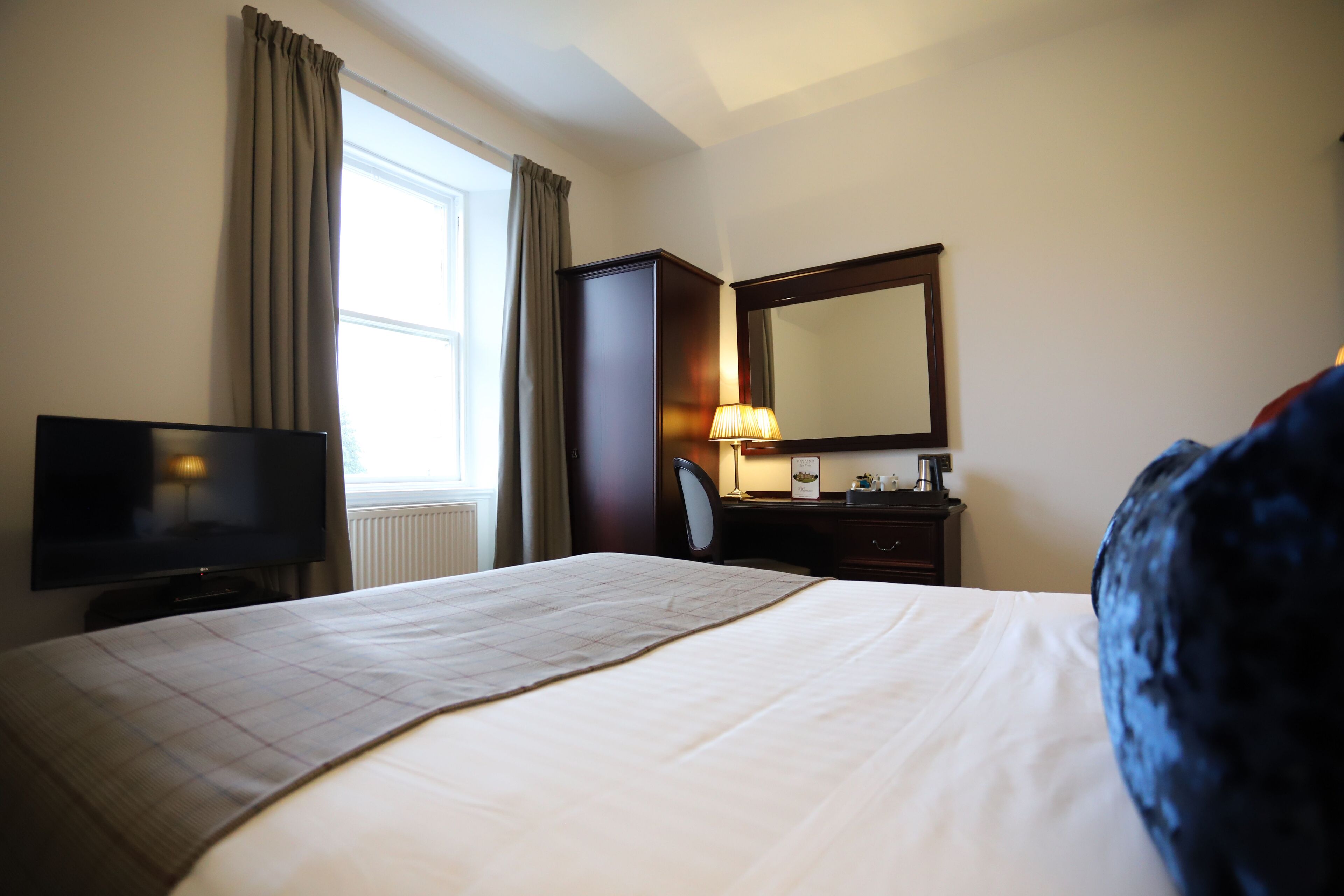 A hotel room with a bed, a television, a desk with a lamp, a mirror, and a window with curtains.