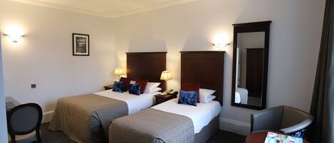 A hotel room with two beds, a desk, a chair, a table with a magazine, and a mirror.
