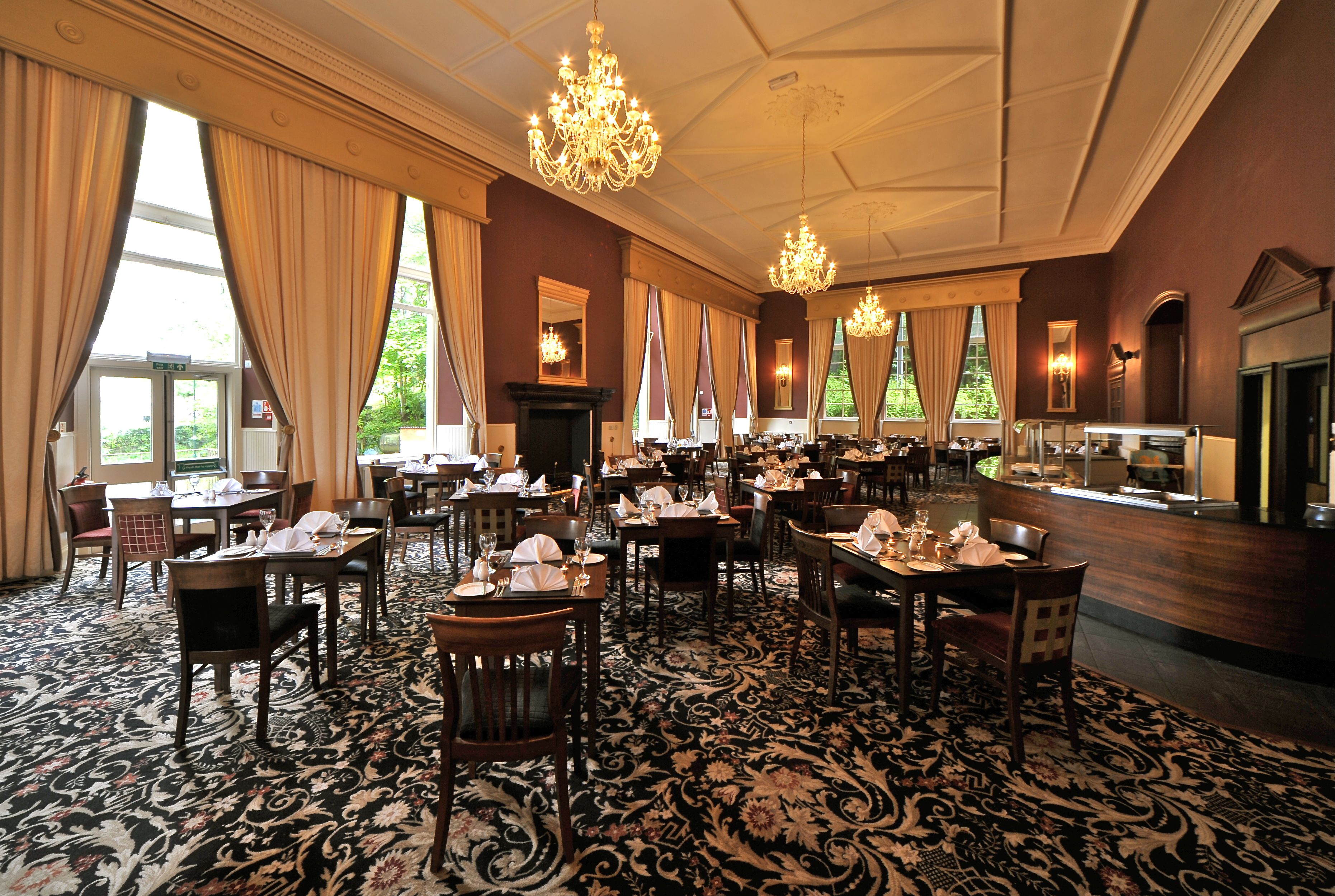 A large dining room with multiple tables set for service, chandeliers, and large windows with curtains.