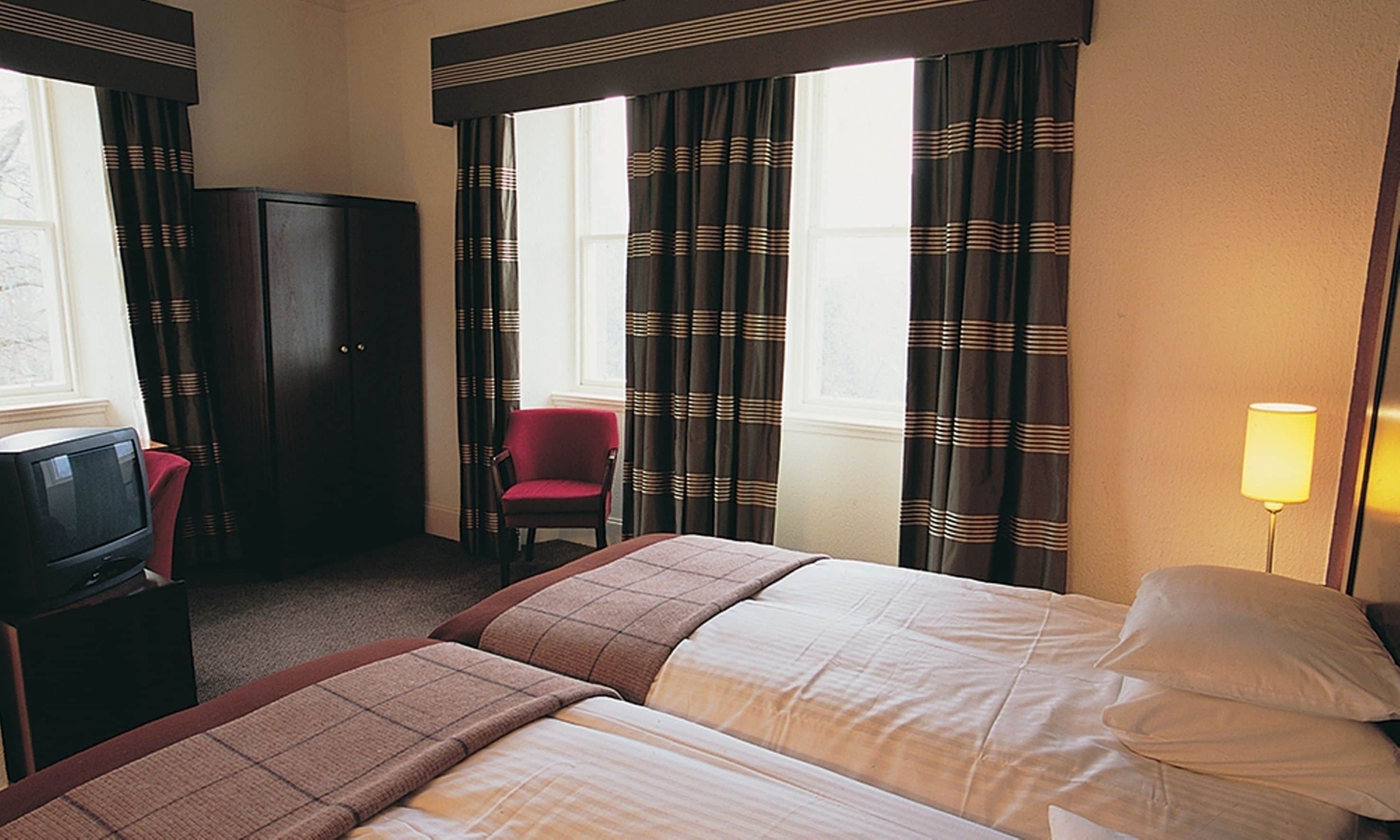 A hotel room with two beds, a desk with a television, a red chair, and a lamp.