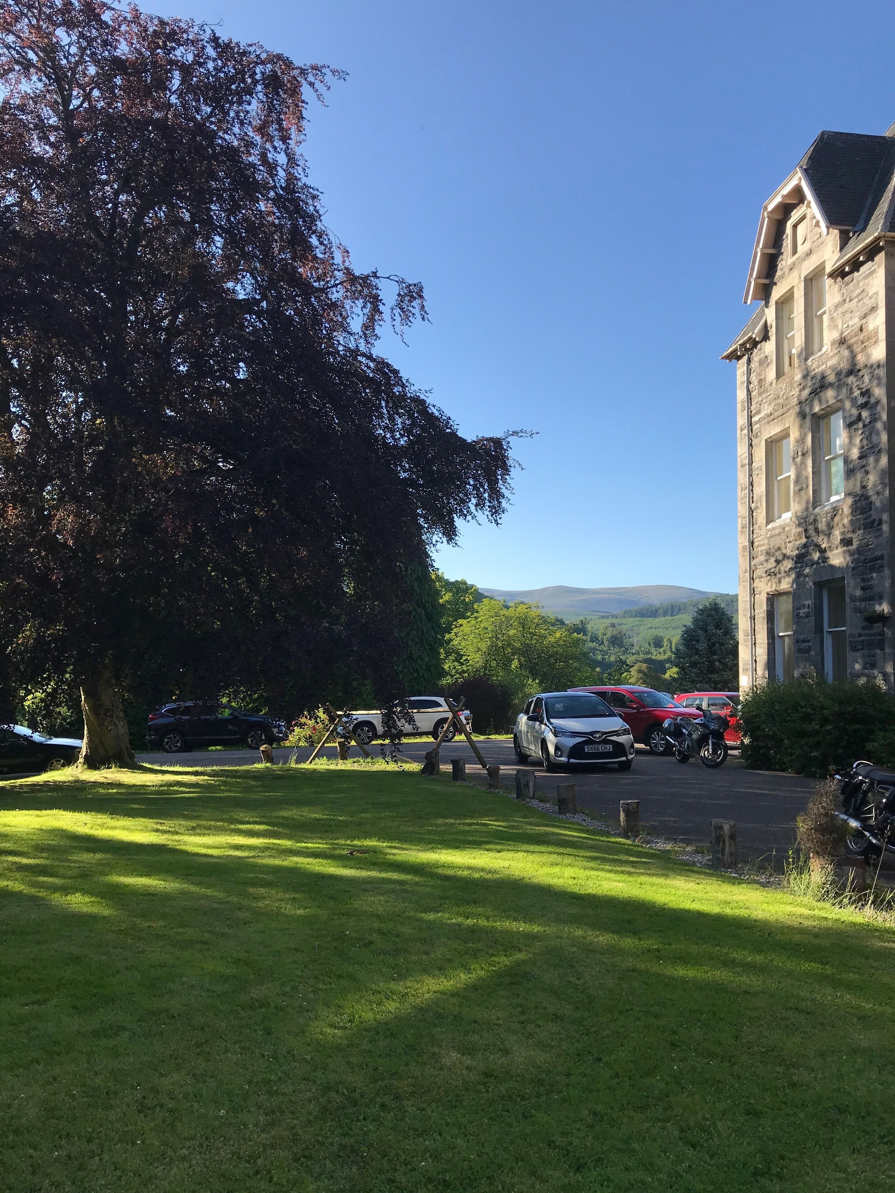 Un aparcamiento con varios coches y una motocicleta, un árbol grande y un edificio de piedra con numerosas ventanas.
