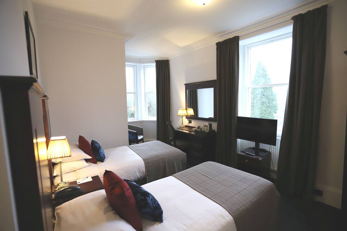 A hotel room with two beds, a desk with a lamp, a television, and a window with curtains.
