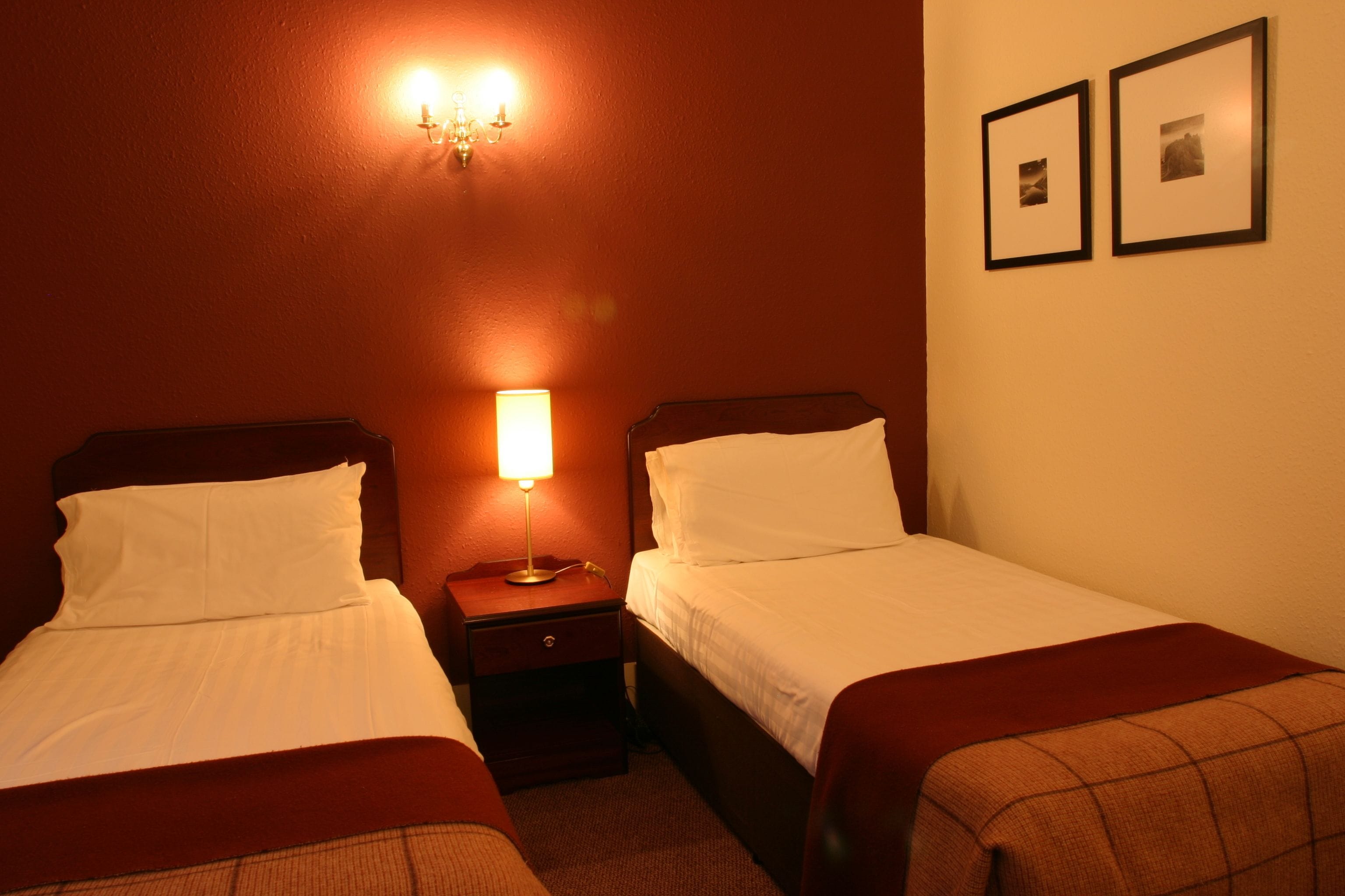 A hotel room with two beds, a nightstand, a wall-mounted lamp, and two framed pictures on the wall.