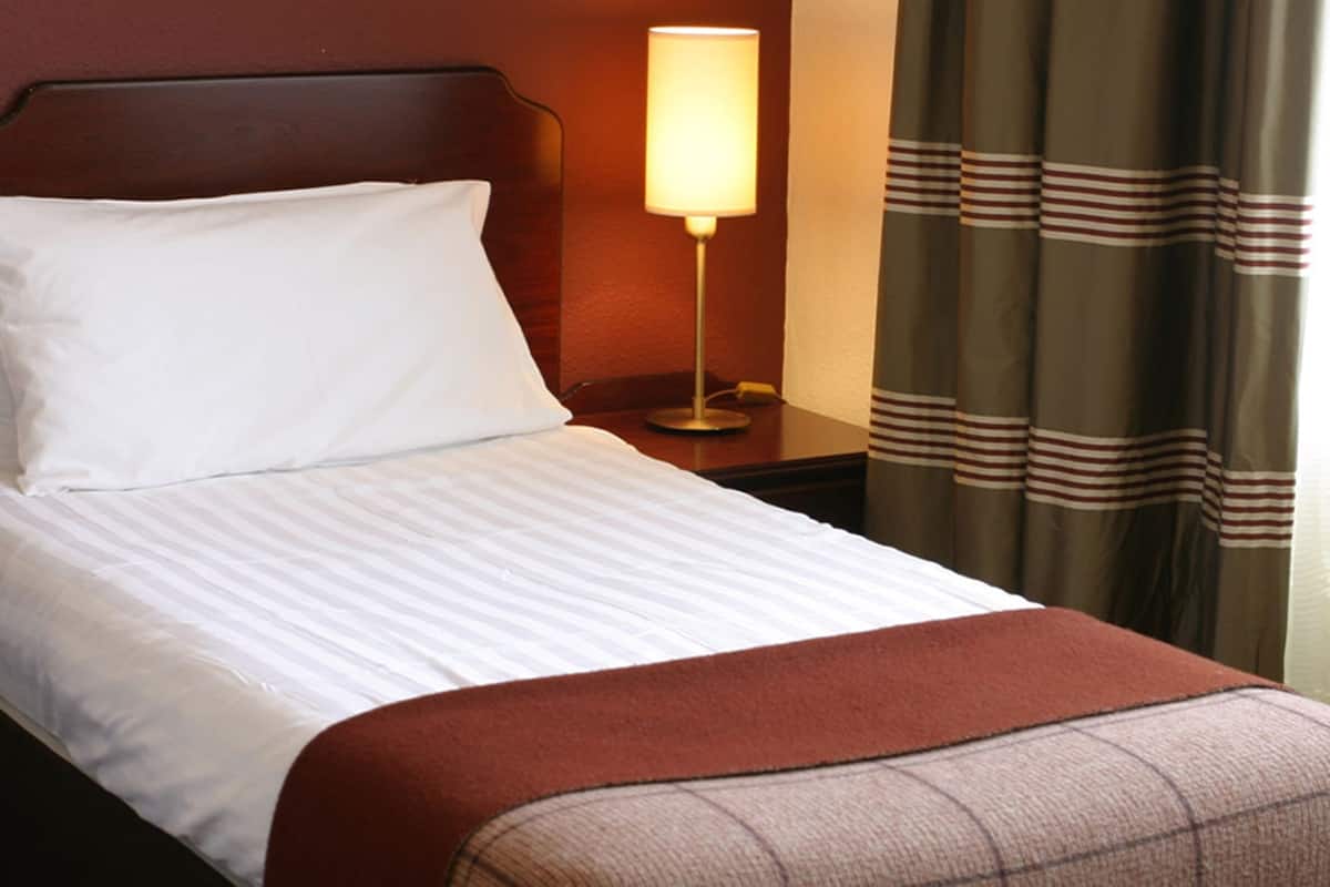 A neatly made bed with white bedding and a brown throw blanket, a bedside lamp, and striped curtains.