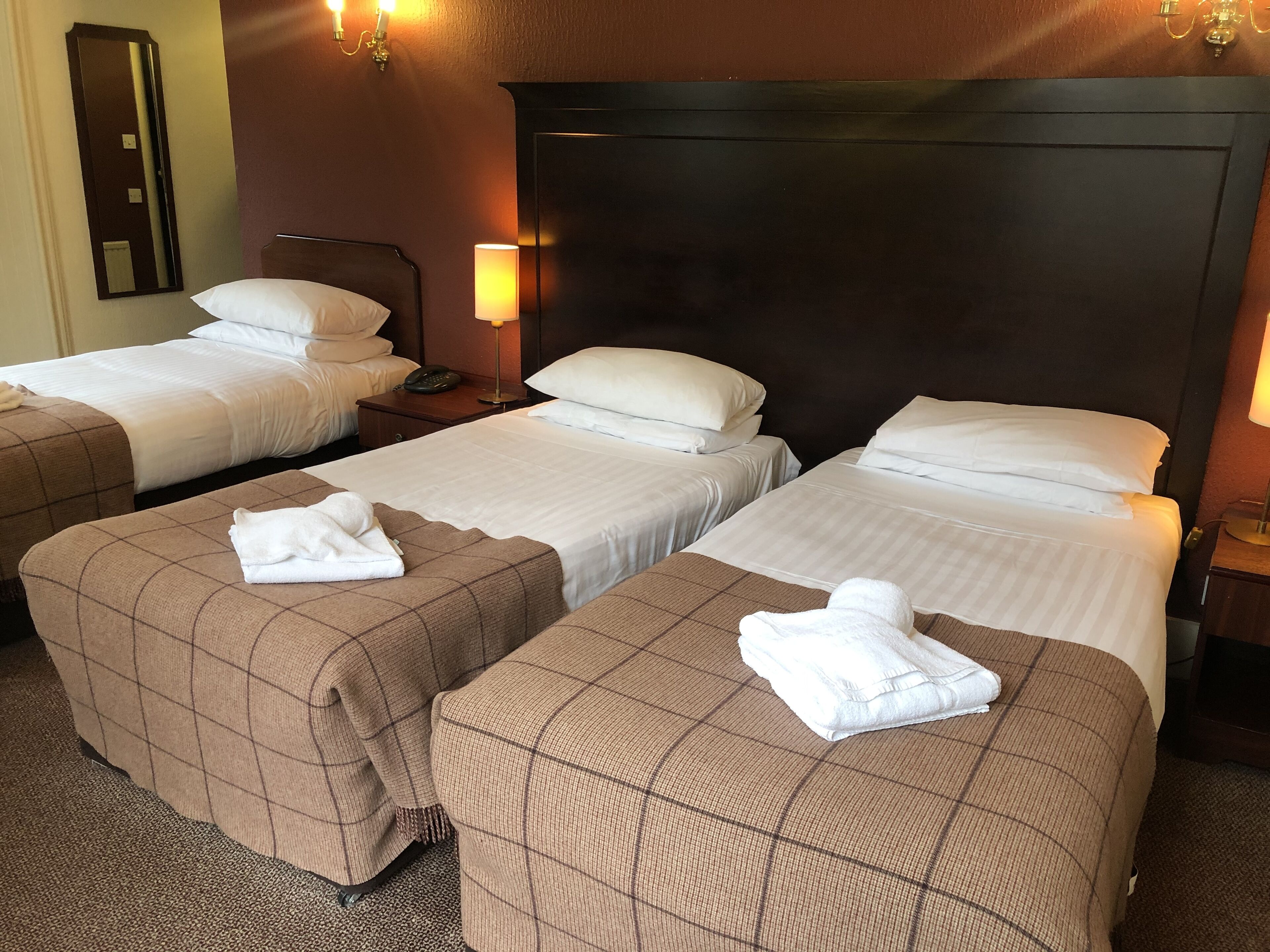 A hotel room with three single beds, each with a brown checkered bedspread and white pillows. There are two bedside lamps, a mirror on the wall, and a telephone on the nightstand.