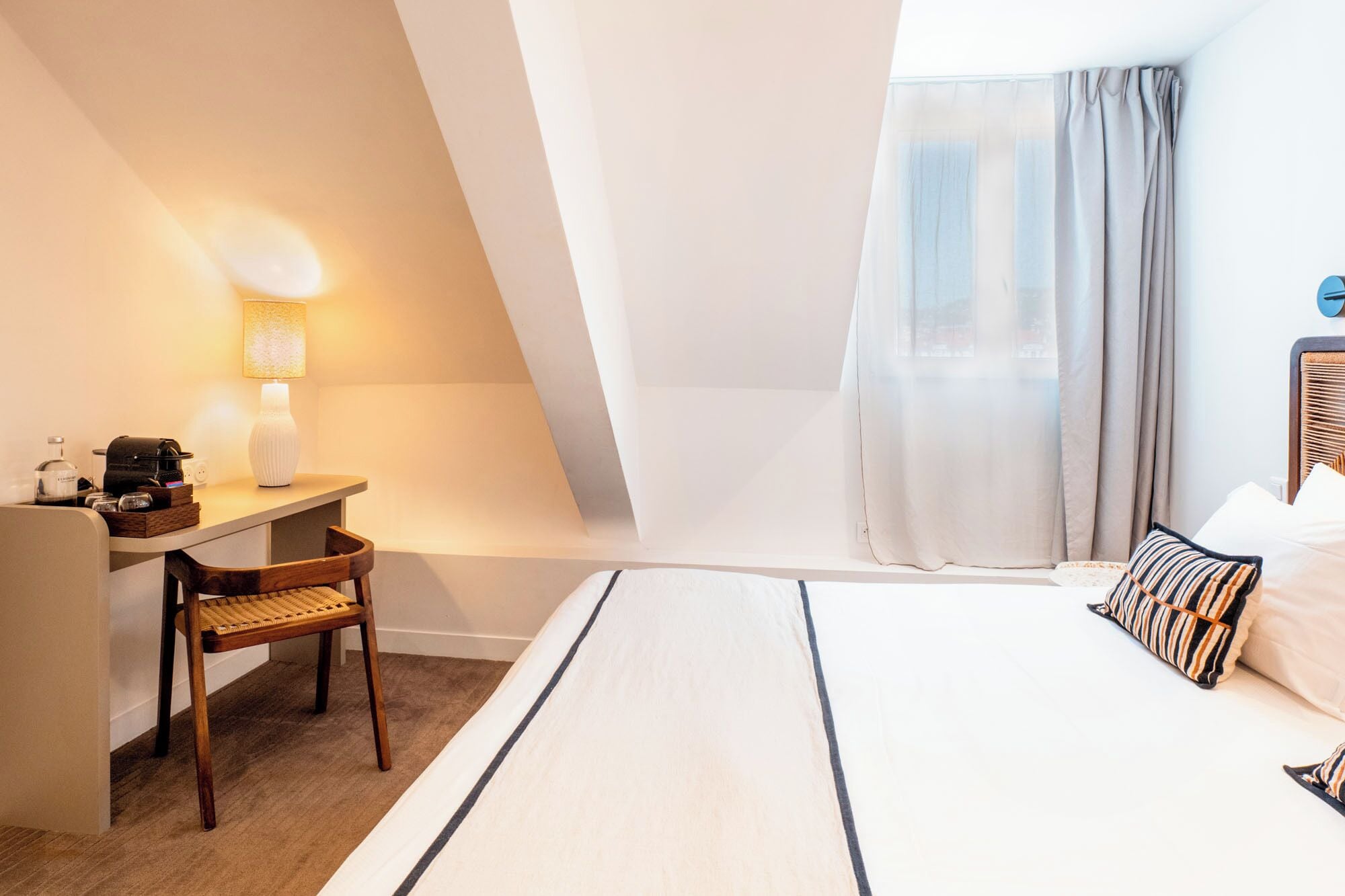 A hotel room with a bed, a desk, a chair, a lamp, and a window with curtains.