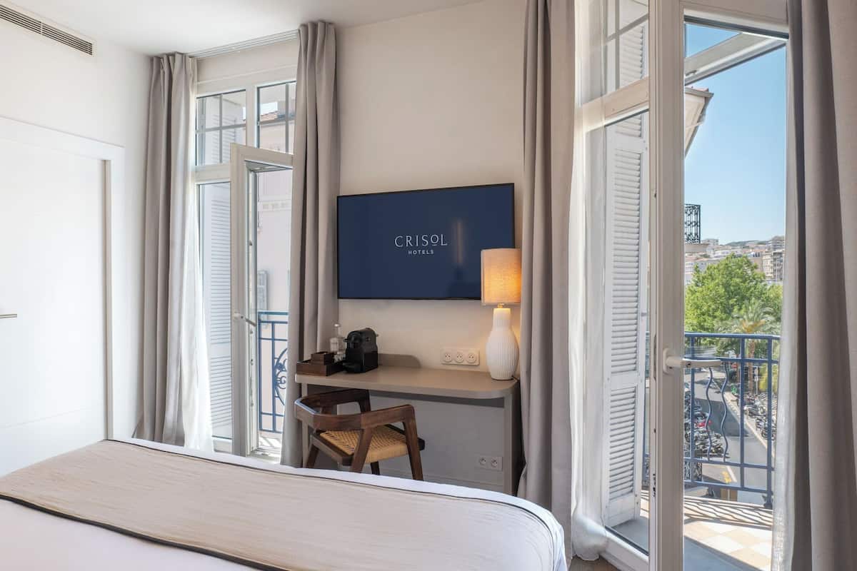 A hotel room with a bed, a desk with a chair, a television, and a balcony with a view of the city.