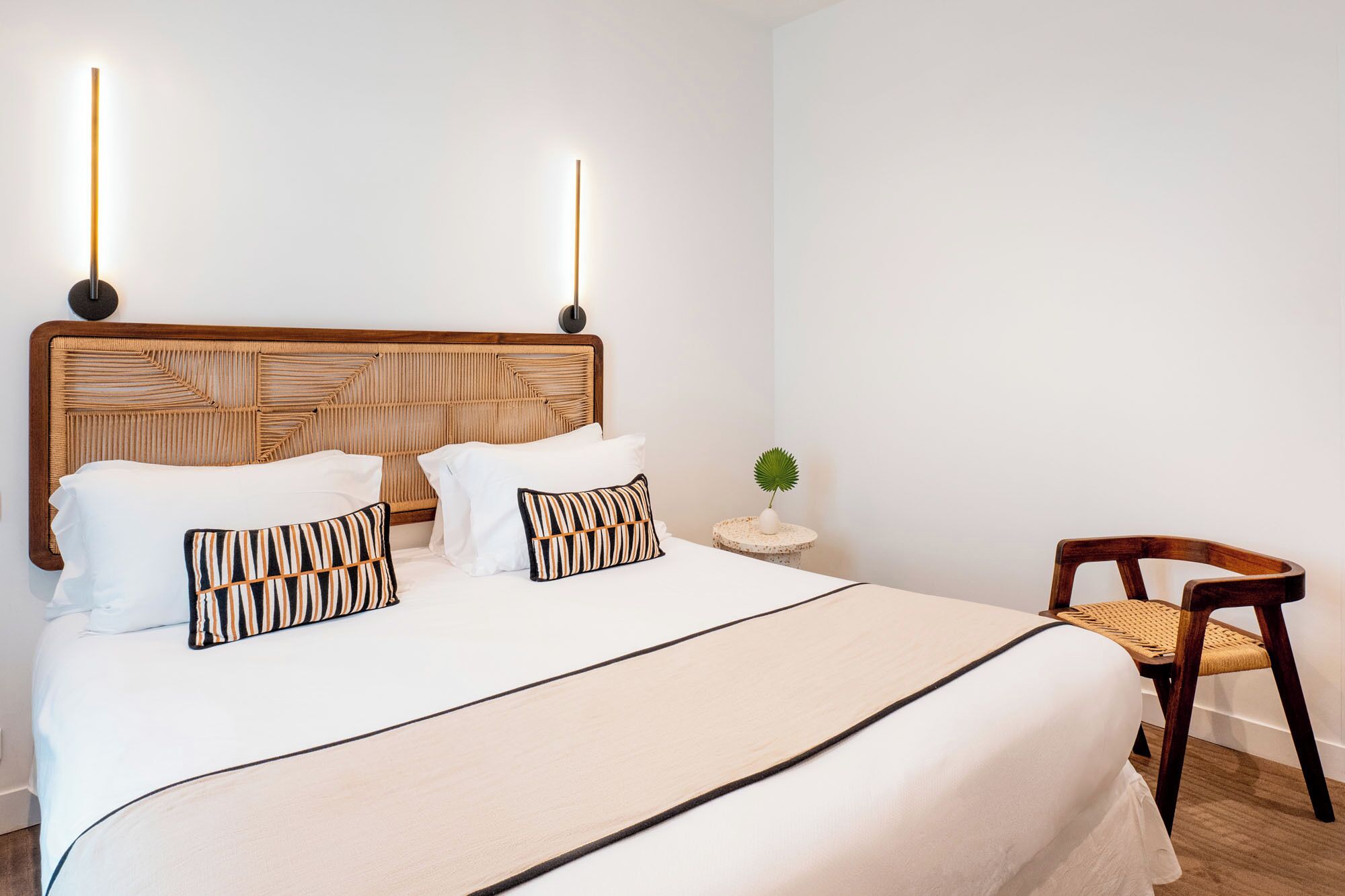 A bed with a wooden headboard, white bedding, and striped pillows. There is a small round table with a plant and a chair with a woven seat.