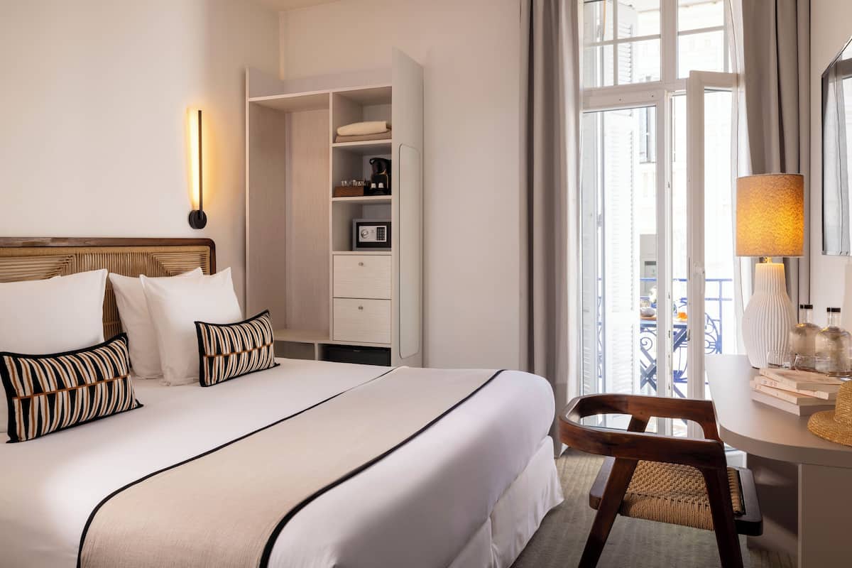 A hotel room with a large bed, a wardrobe, a desk with a lamp, and a view of the outside.