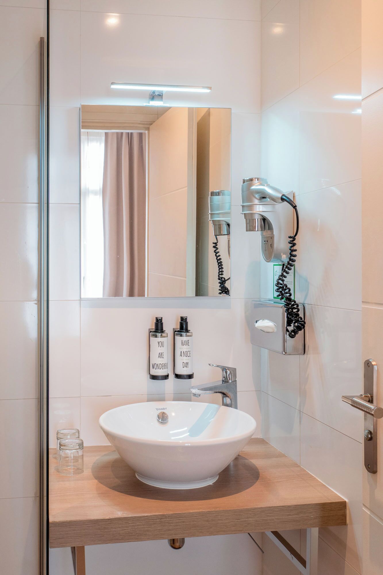 Une salle de bain moderne avec un lavabo blanc, un miroir, un sèche-cheveux et deux bouteilles ornées de messages inspirants.