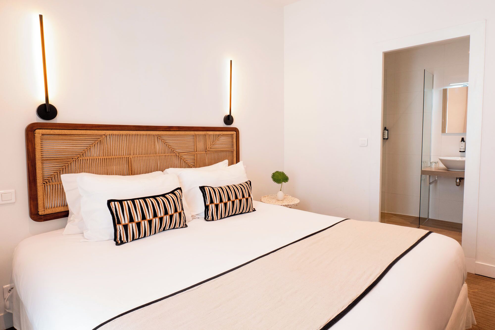 A hotel room with a large bed, a wooden headboard, bedside lamps, a small round table with a plant, and a bathroom visible through an open door.