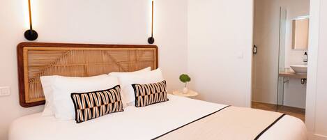 A hotel room with a large bed, a wooden headboard, bedside lamps, a small round table with a plant, and a bathroom visible through an open door.
