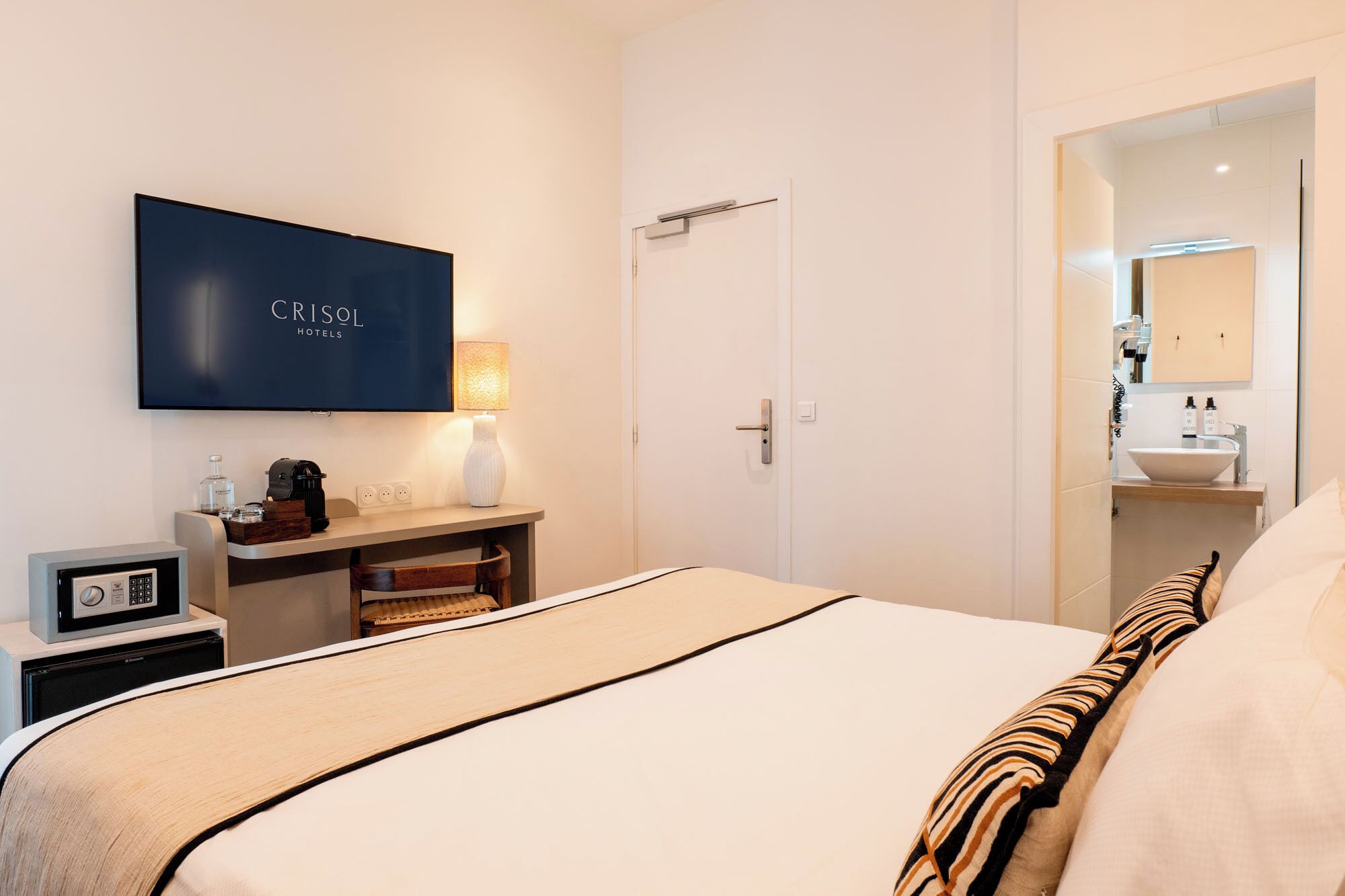 Una habitación de hotel con cama, televisor montado en la pared, mesita de noche con lámpara y puerta que da a un baño.