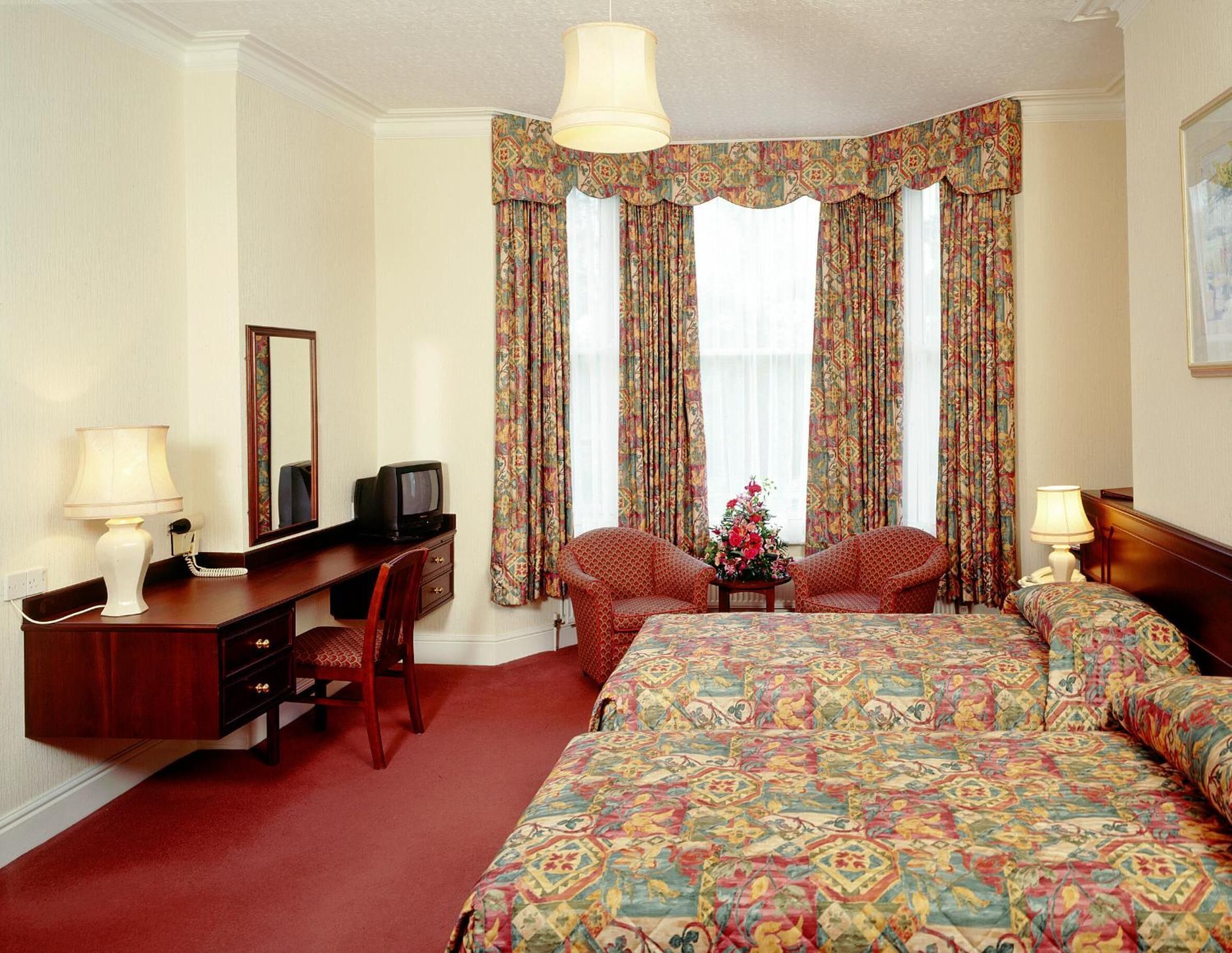 A hotel room with a large bed, two armchairs, a desk with a telephone and television, and a window with patterned curtains.