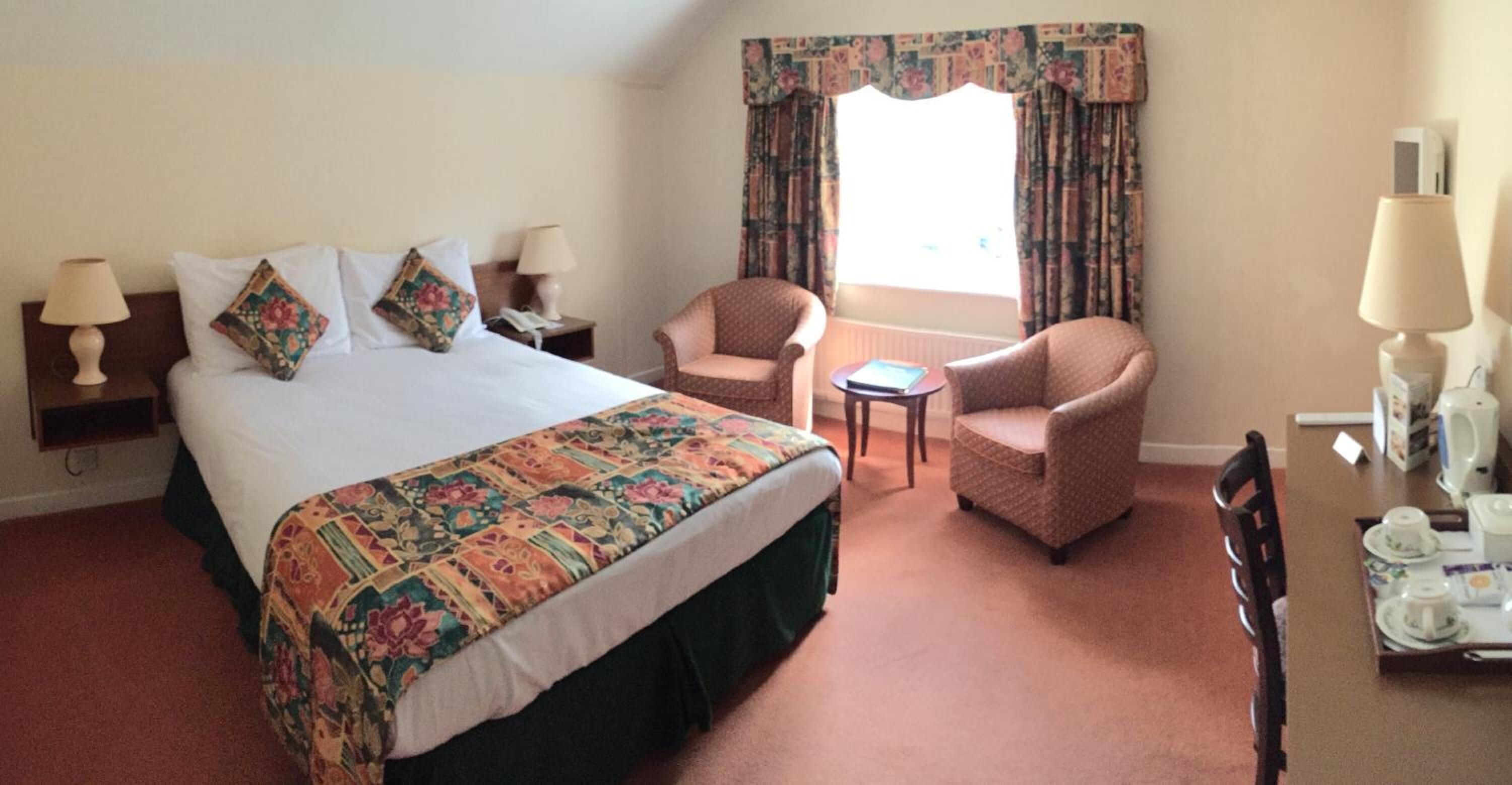 A hotel room with a large bed, two chairs, a small table, and a desk with a kettle and cups.