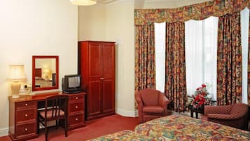 A hotel room with a large bed, a desk with a television, two armchairs, a wardrobe, and a mirror.
