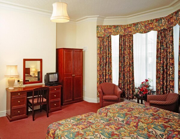 Une chambre d’hôtel avec un grand lit, un bureau avec une télévision, deux fauteuils, une armoire et un miroir.