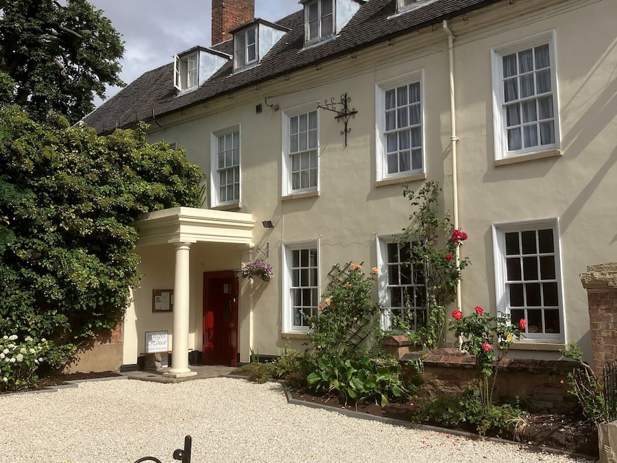 A traditional building with a gravel driveway, a gated entrance, and a flower bed with red flowers.