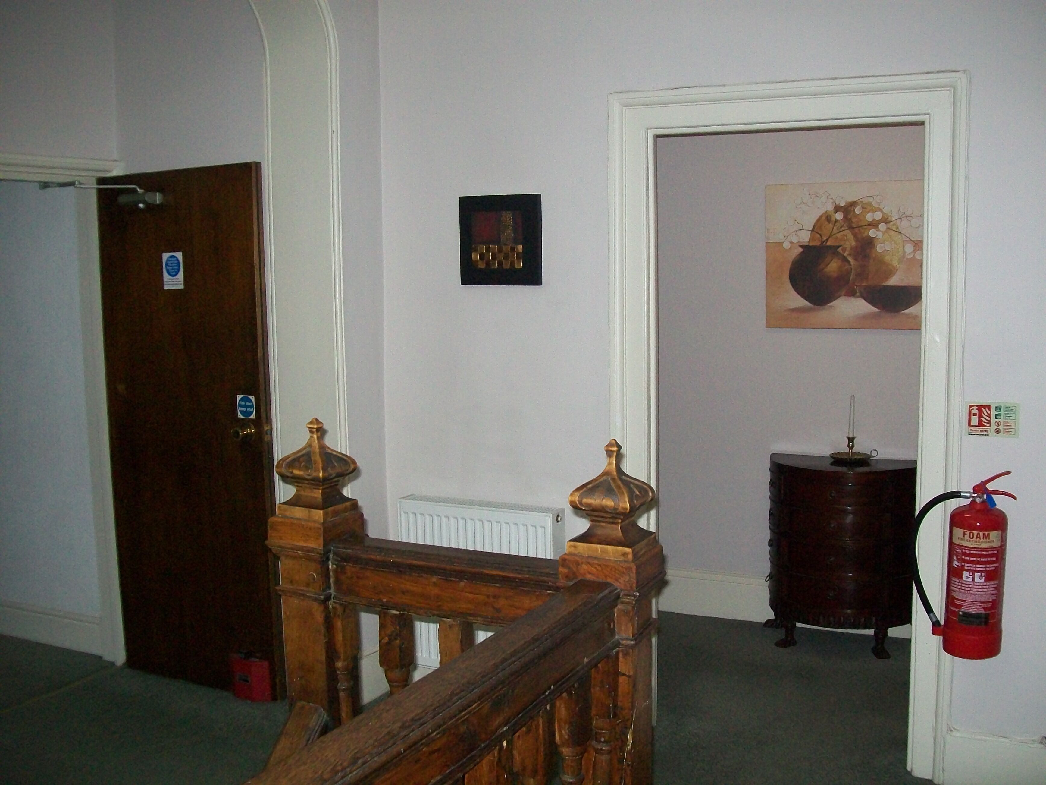 A hallway with a wooden staircase, a door with a sign, a framed picture on the wall, and a fire extinguisher mounted on the wall.