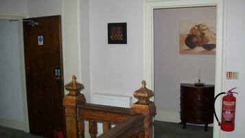 A hallway with a wooden staircase, a door with a sign, a framed picture on the wall, and a fire extinguisher mounted on the wall.