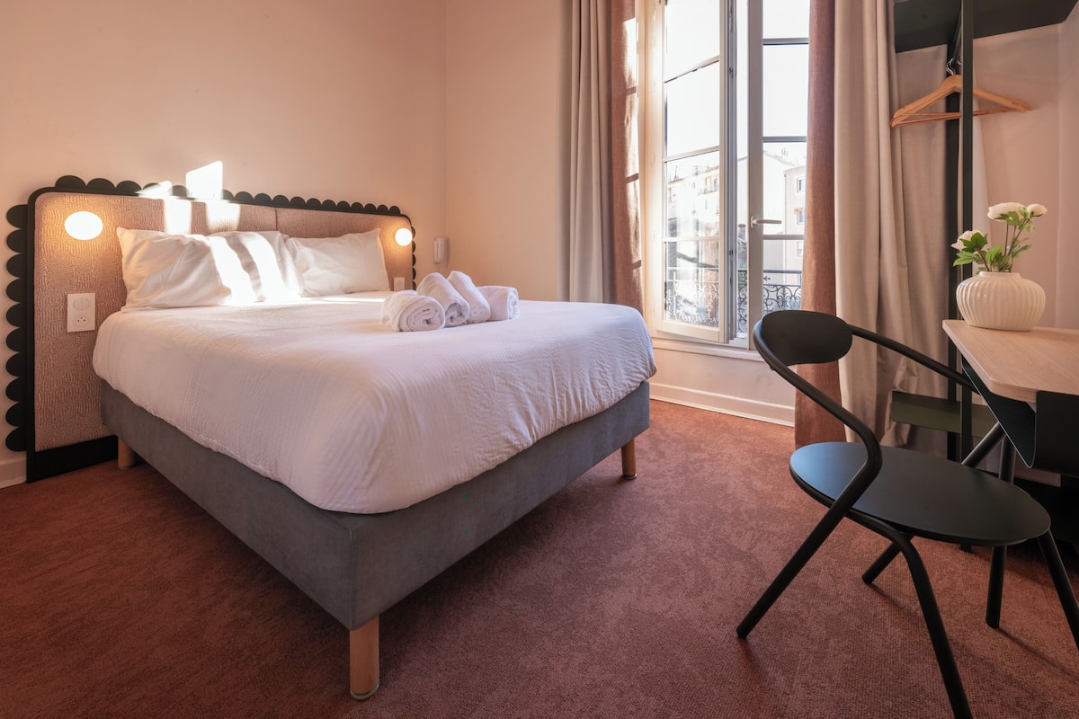 A neatly made bed with white linens and a headboard with circular lights, a small table with a vase of flowers, and a chair.