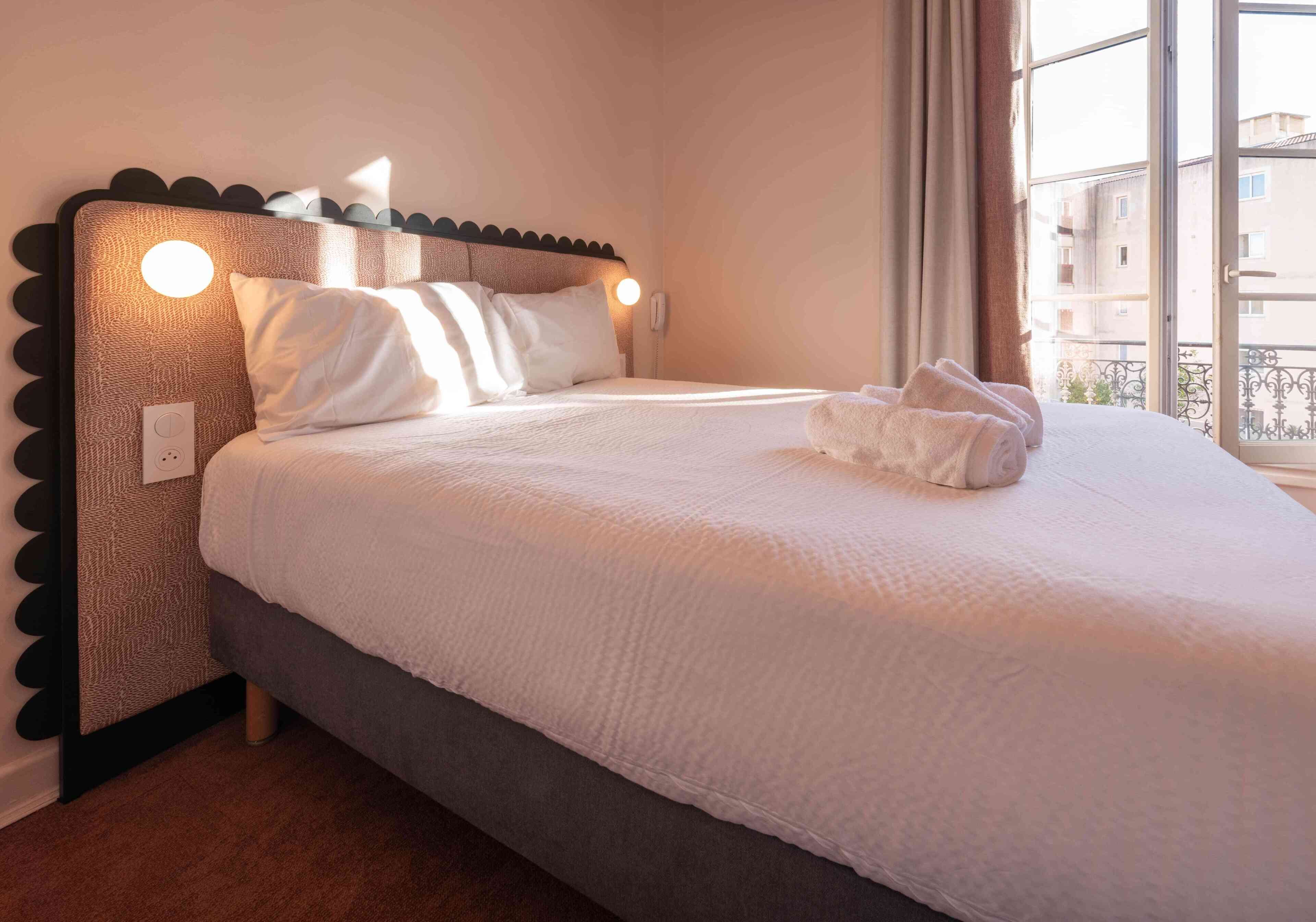 A neatly made bed with white linens, a headboard with circular light fixtures, and a window with a view of buildings.