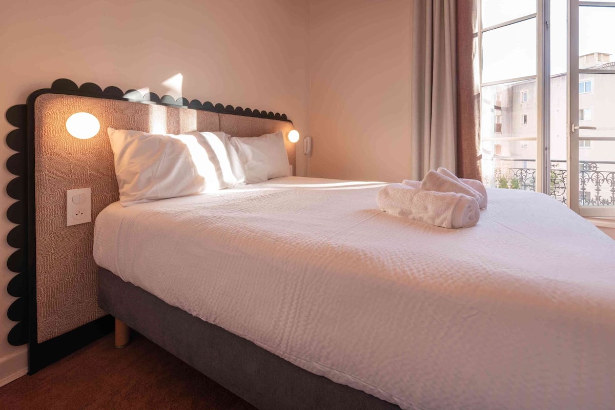 A neatly made bed with white linens, a headboard with circular light fixtures, and a window with a view of buildings.