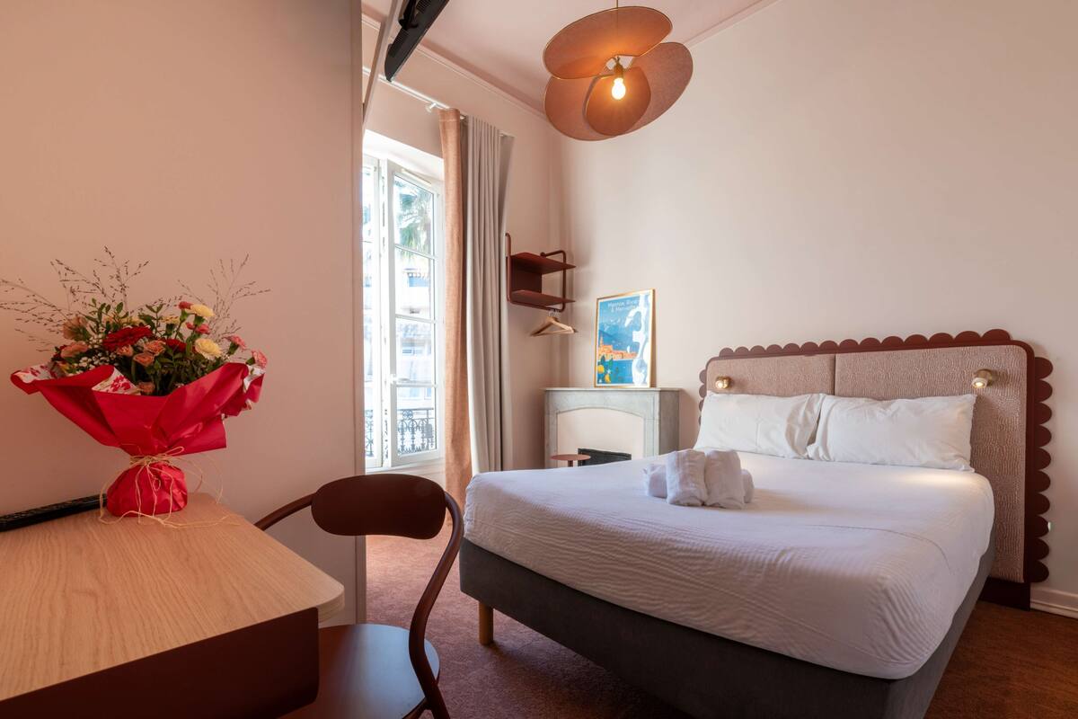 A hotel room with a bed, a desk with a vase of flowers, a chair, a bedside table, a lamp, and a window with curtains.