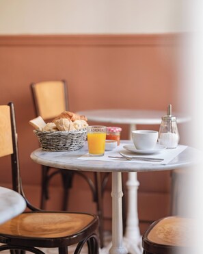 A dining table with a basket of bread, a glass of orange juice, a cup, and a salt shaker.